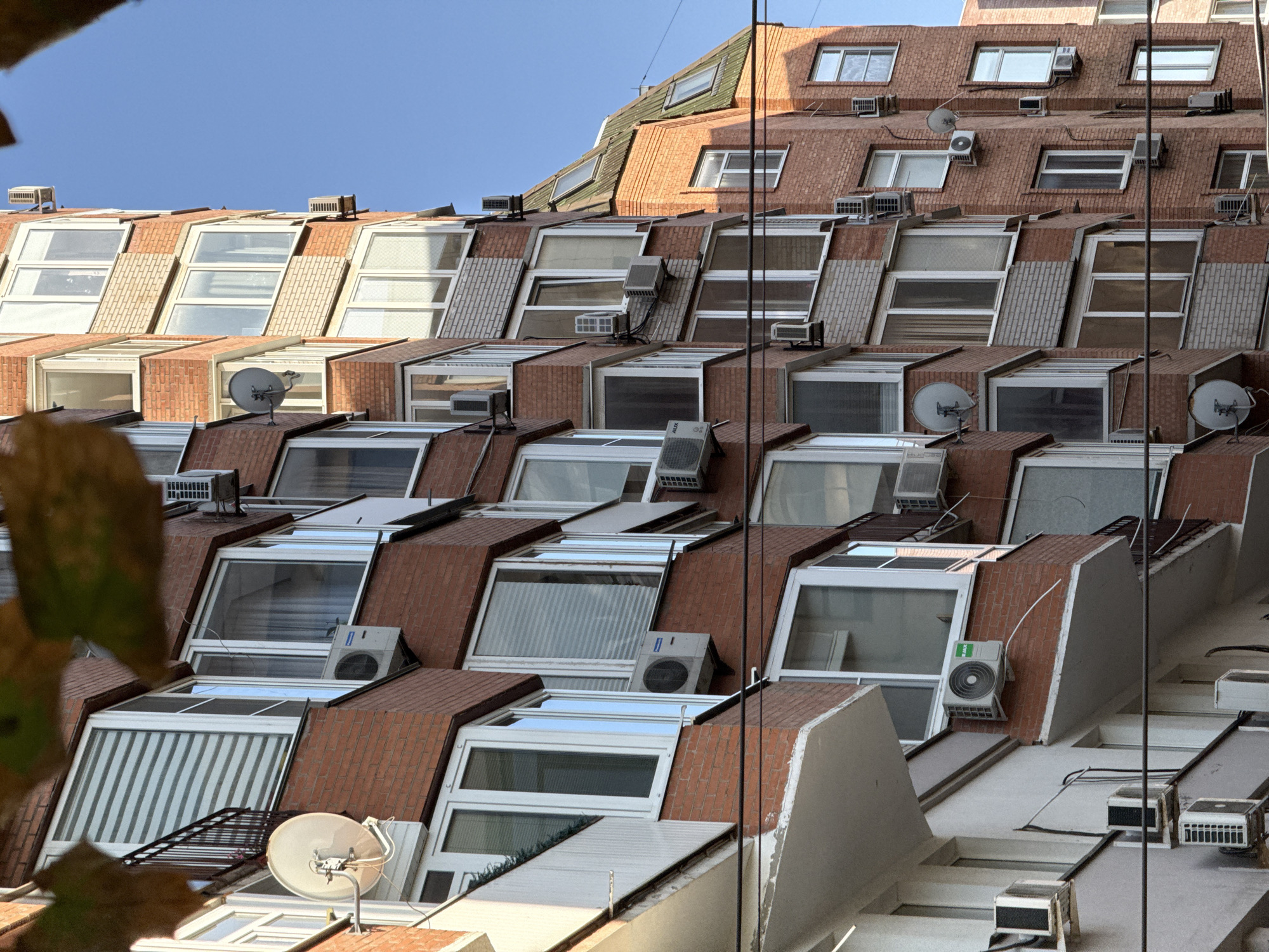 The image shows a close-up, low-angle view of a multi-story brick apartment building in Rostov-on-Don, Russia. The building features numerous windows and balconies, many of which are equipped with air conditioning units. Several satellite dishes are also visible mounted on the exterior walls. The architecture suggests a post-Soviet era construction. The sky is clear and blue, indicating a sunny day. Some autumn leaves are partially obscuring the top of the frame. There are no people visible in the image. The scene captures a common urban residential environment.