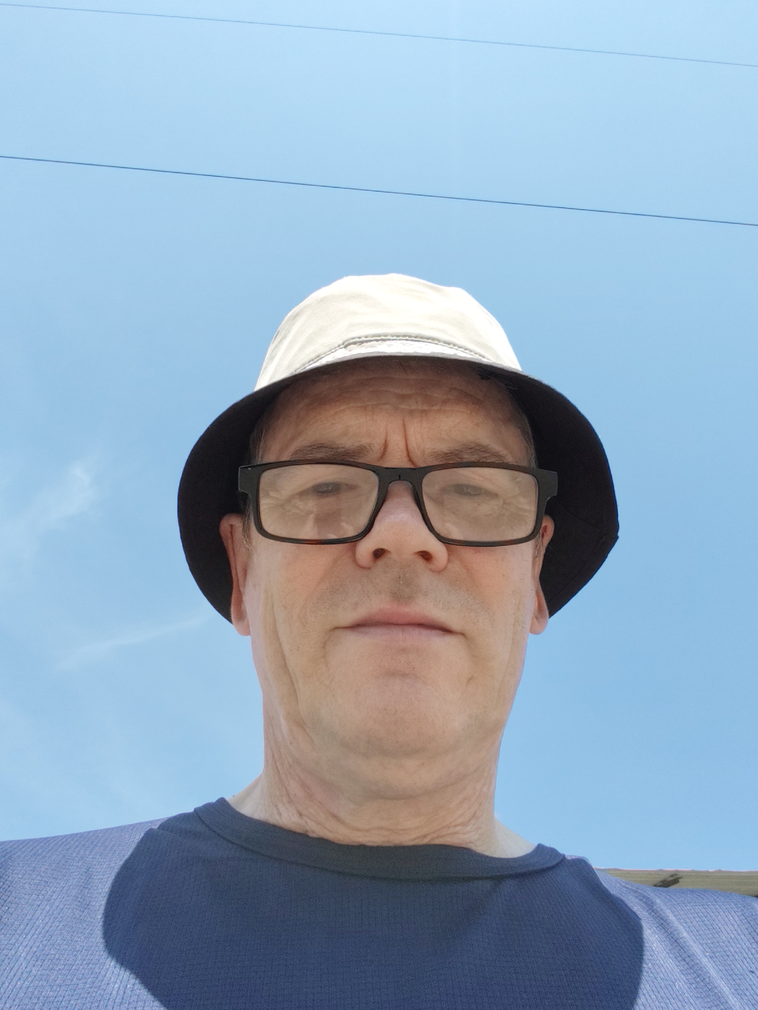 A low-angle, close-up outdoor selfie shows a Caucasian man wearing glasses and a bucket hat against a clear blue sky. He is looking directly at the camera and is wearing a dark blue t-shirt. The image appears to have been taken during the daytime. There is no indication of the city of Komsomolsk-on-Amur, Russia.