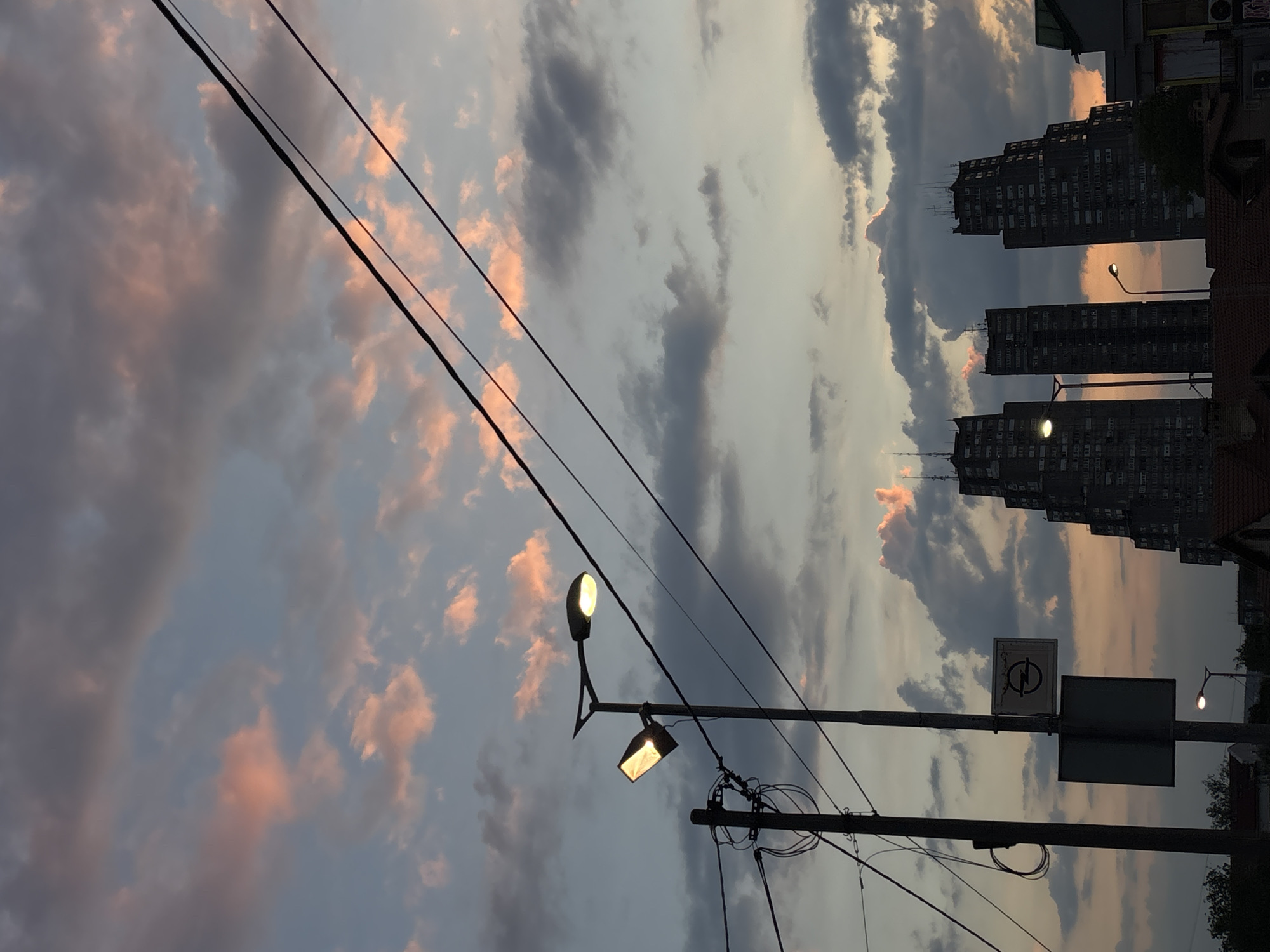 The photograph captures a street scene in Belgrade, Serbia, at dusk. Three buildings, likely apartment blocks, rise in the background. Clouds with pink highlights dominate the sky. Several streetlights and utility poles stand along the street. A sign post is also visible.