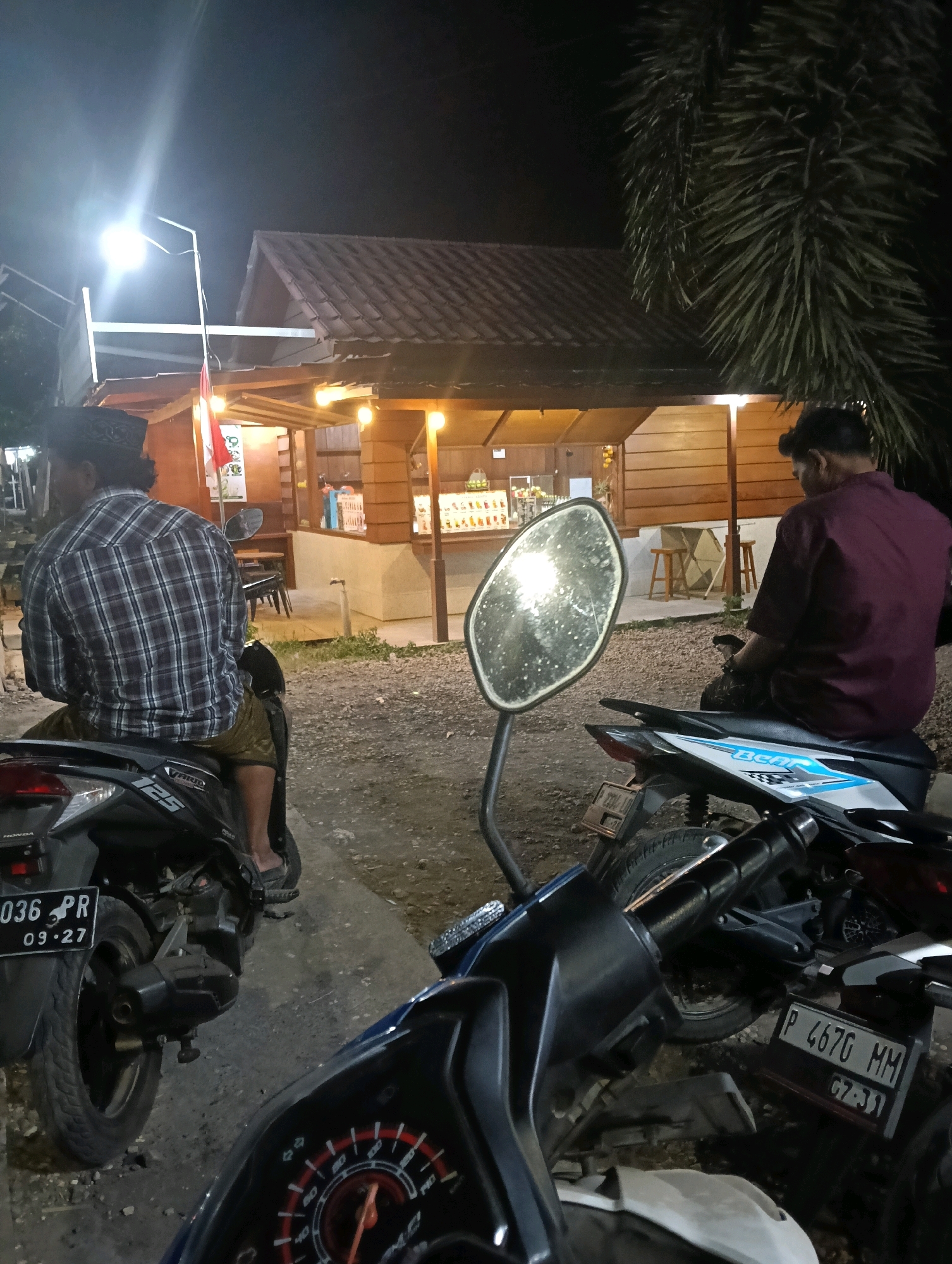 The image depicts a nighttime scene outside a small wooden building, possibly a food stall or shop, illuminated by artificial lights. The building has a tiled roof and wooden walls, with a counter area visible inside showcasing drinks or food items. Two men are seated on motorcycles in the foreground. The man on the left is wearing a plaid shirt and a black skullcap, facing away from the camera. The man on the right is wearing a maroon shirt and is also facing away from the camera. The motorcycles are parked on a gravelly surface. In the background, a tall palm tree is visible. A bright light fixture is mounted on a pole, illuminating the area. The overall atmosphere suggests a casual gathering or stop at a local establishment in Grenden, Indonesia.