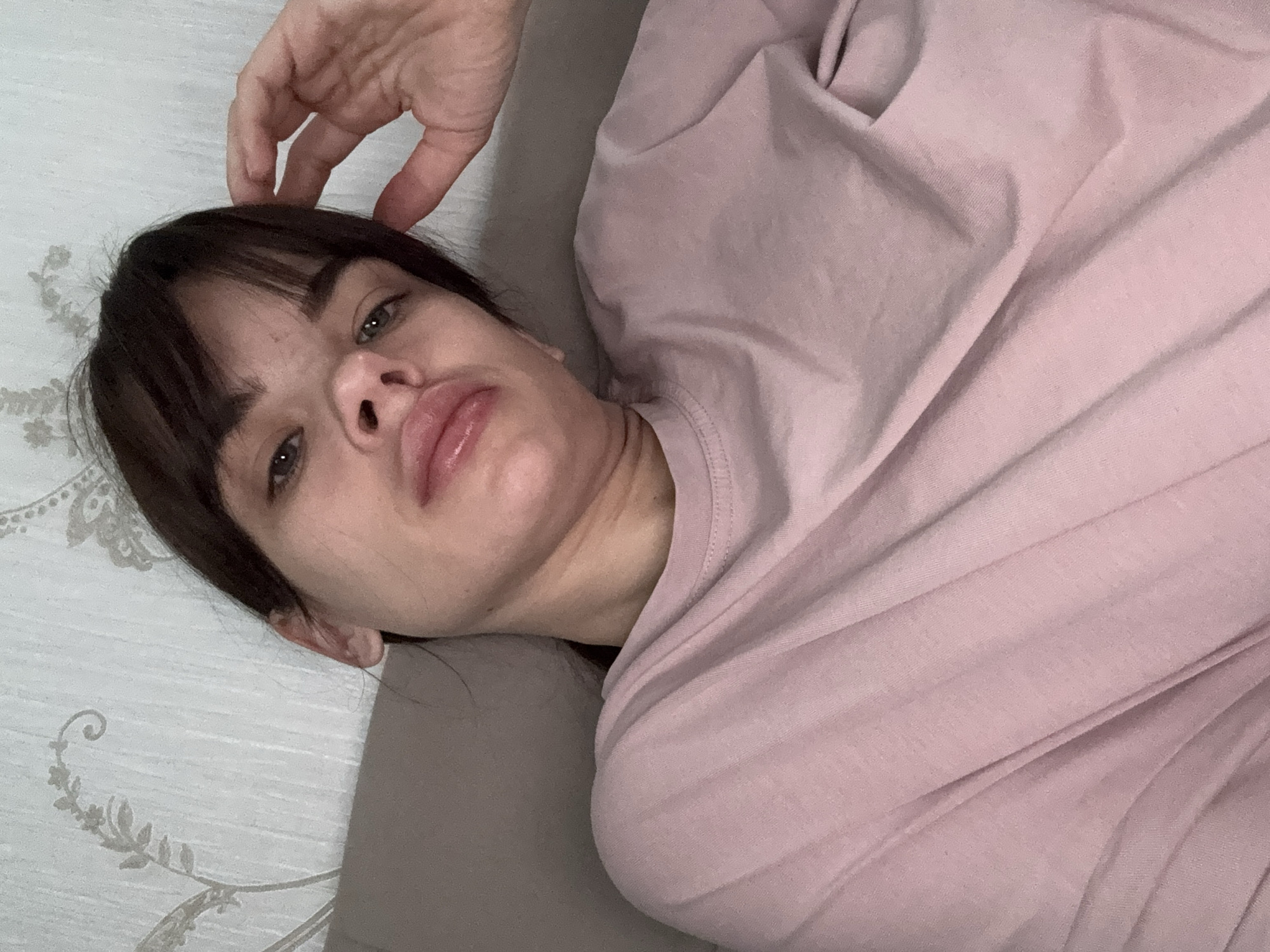 A woman lies on a patterned light-colored surface (possibly a bed), wearing a pink long-sleeved shirt. Her head is tilted, and she is looking up. A hand is near her head, possibly touching it. The location cannot be determined from the image, and there is no indication that the photo was taken in Rostov-on-Don, Russia.