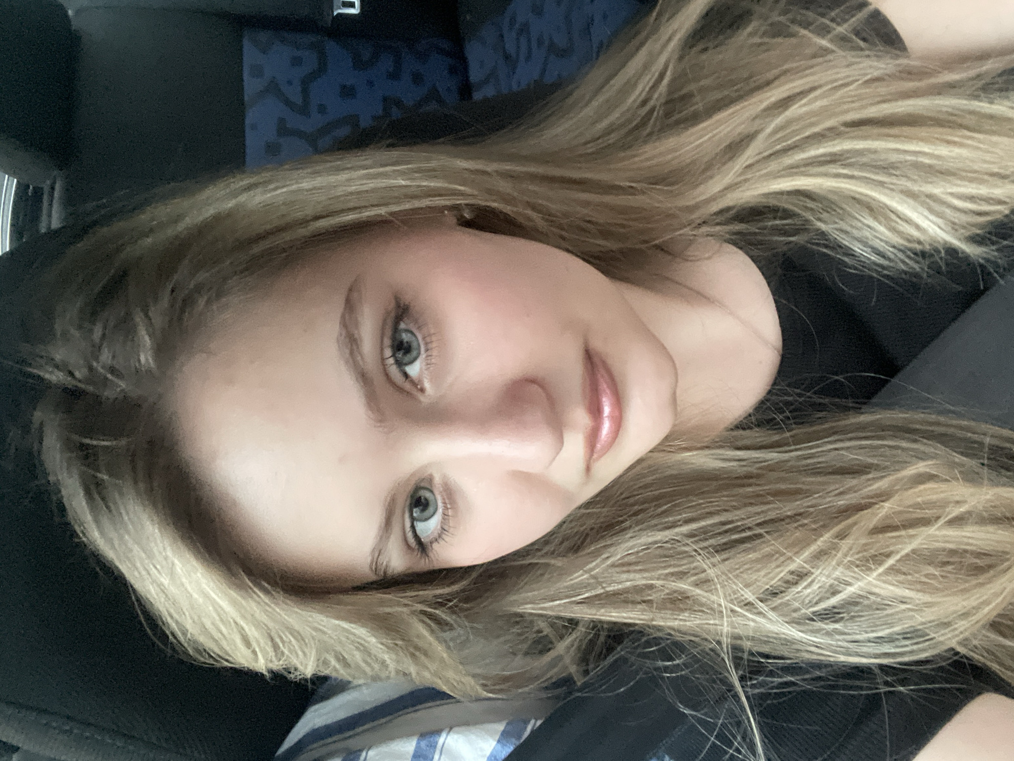 A young woman with blonde hair and blue eyes is shown from the chest up. She is wearing a black shirt and sitting in a car, indicated by the car seat behind her. Her hair is long and flows down past her shoulders. The lighting suggests it is daytime.