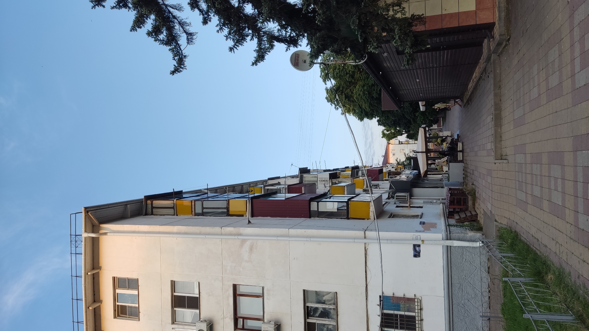 The image shows a street scene in Krasnodar, Russia, with a multi-story apartment building on the left and a dark wooden building on the right. The sky is clear blue. The apartment building has several balconies with yellow and red trim, and air conditioning units are visible. A paved sidewalk runs between the buildings, with some outdoor seating under umbrellas in front of a business on the right. A satellite dish is mounted on the wall of the wooden building. A pine tree extends into the frame from the upper right.