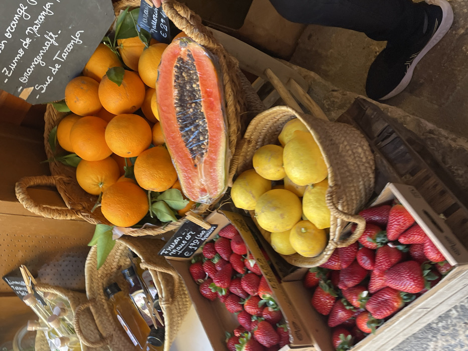 This image depicts a market stall or shop entrance in Valldemossa, Spain, showcasing fresh produce. The scene is dominated by multiple containers of fruit. A large, woven wicker basket in the center-right is filled with numerous bright orange oranges, some retaining green leaves. Resting horizontally atop these oranges is a large papaya, cut lengthwise to reveal its dark seeds, and wrapped in clear plastic. Above the oranges, a black chalkboard sign lists various terms for "orange juice" in multiple languages. To the lower left of the oranges, a smaller woven wicker basket contains a pile of bright yellow lemons. In the foreground and to the left, two wooden crates are filled with ripe red strawberries. A small black chalkboard sign on the upper strawberry crate reads "Fresas Mallorca €1.90." In the upper left background, another wicker basket holds several small, clear glass bottles with wooden stoppers, likely containing liquids such as oils or vinegars. The produce is displayed on rustic wooden planks or crates, which rest on a light-colored, uneven stone or tiled floor. In the lower right corner, a person's left leg, clad in dark pants and a black athletic shoe with a white sole, is visible, standing adjacent to the produce display. A hand is partially visible touching a chalkboard sign on the right.