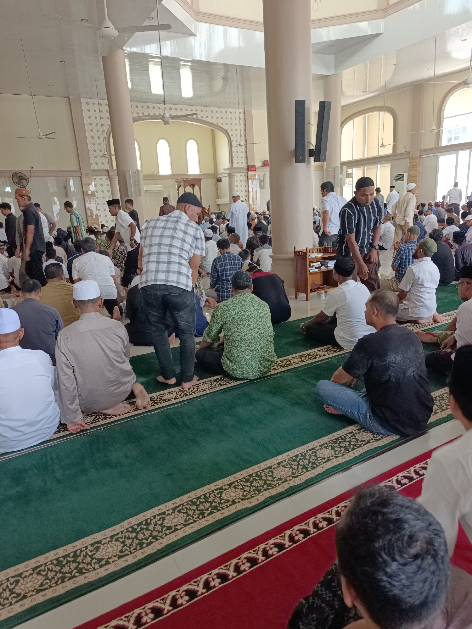 The image shows a large group of men gathered inside a mosque, likely for prayer. The setting appears to be the city of Mesjid Reuntoh, Indonesia. The mosque features high ceilings with white fans, large arched windows, and beige walls with decorative patterns. Pillars are also visible throughout the space. The floor is covered with green carpet, bordered by a red carpet and ornate rug patterns. The men are dressed in various attire, including traditional Islamic clothing like thobes and peplos, as well as casual wear like t-shirts and jeans. Many are seated on the floor, facing what would be the direction of prayer (qibla). Some men are standing or walking, indicating the ongoing activity within the mosque. A wooden shelf unit containing books is visible against one of the pillars. The overall scene depicts a communal gathering for religious purposes within a mosque environment.