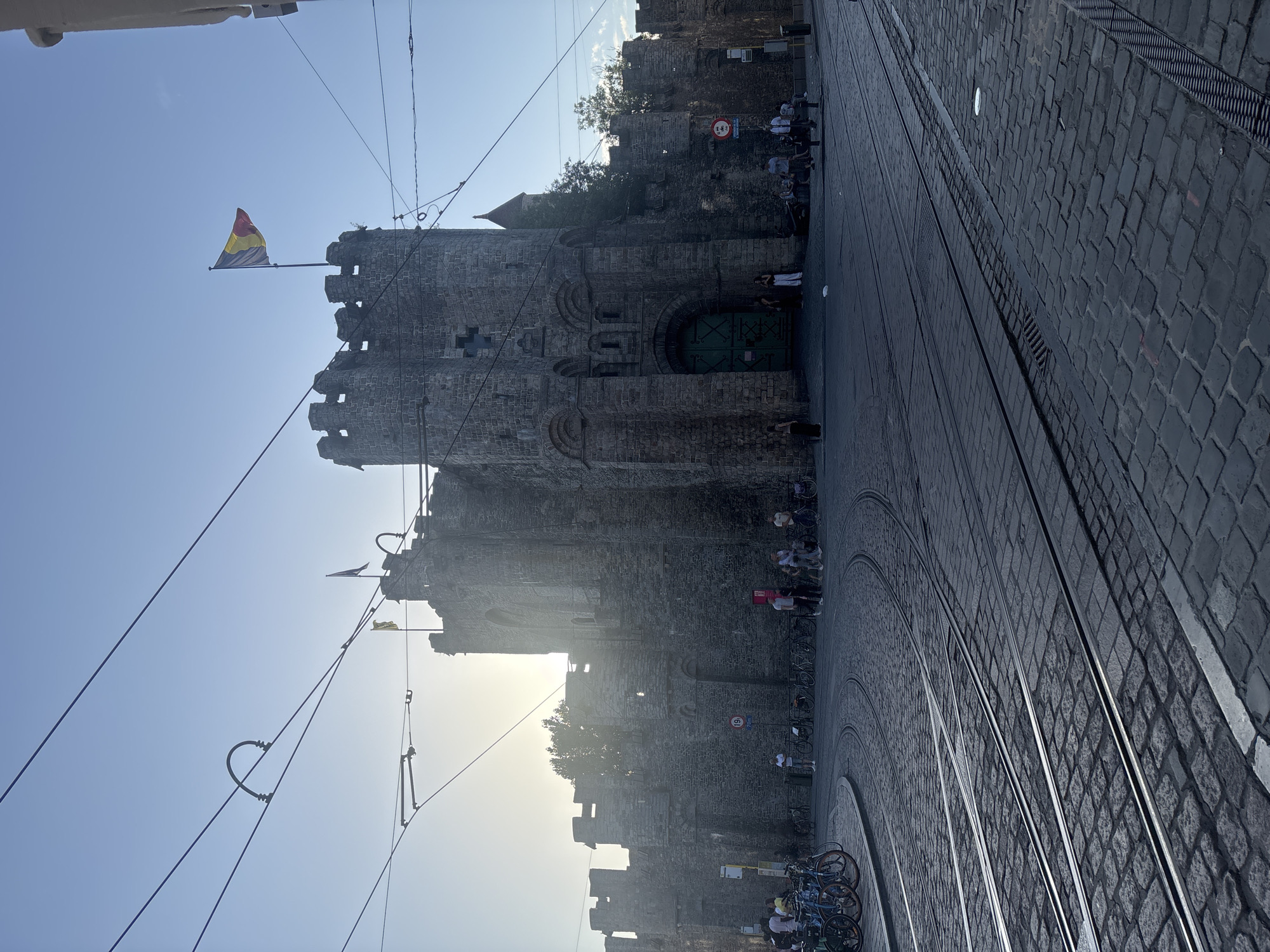 The image depicts a historic stone castle in Ghent, Belgium, with a Belgian flag flying from its ramparts. The castle features a large, arched green entrance gate and is surrounded by a cobbled street with tram tracks. Several people are visible near the entrance, some standing and others sitting on benches. A group of bicycles is parked on the left side of the frame, and more bicycles are seen further down the street. Overhead, electrical wires for trams crisscross the sky, with the sun setting behind the castle, casting long shadows and creating a bright sky. The overall scene suggests a public space in a European city, likely a tourist attraction, during the late afternoon or early evening.