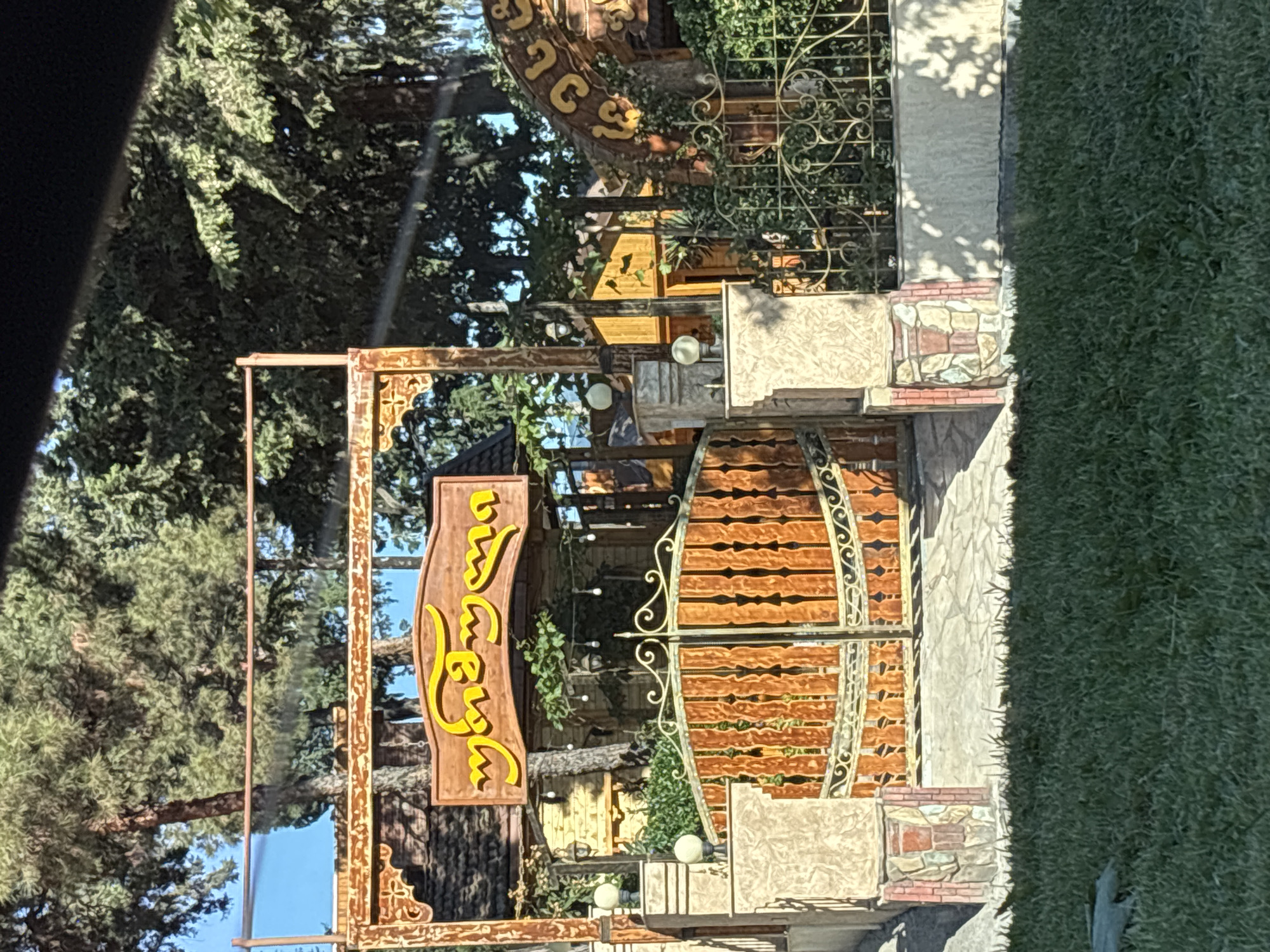 The image shows the entrance to a establishment. A wooden gate with ornate metalwork is visible, flanked by stone pillars topped with spherical lamps. A wooden sign with yellow Georgian script is mounted above the gate. Behind the gate, a building with a thatched roof and wooden walls is partially visible, along with string lights. The entrance is framed by trees and a decorative archway with more Georgian script. The foreground is a patch of green grass. The overall impression is that of an outdoor dining or entertainment venue, possibly in a suburban or park-like setting within Tbilisi, Georgia, given the script on the signage.