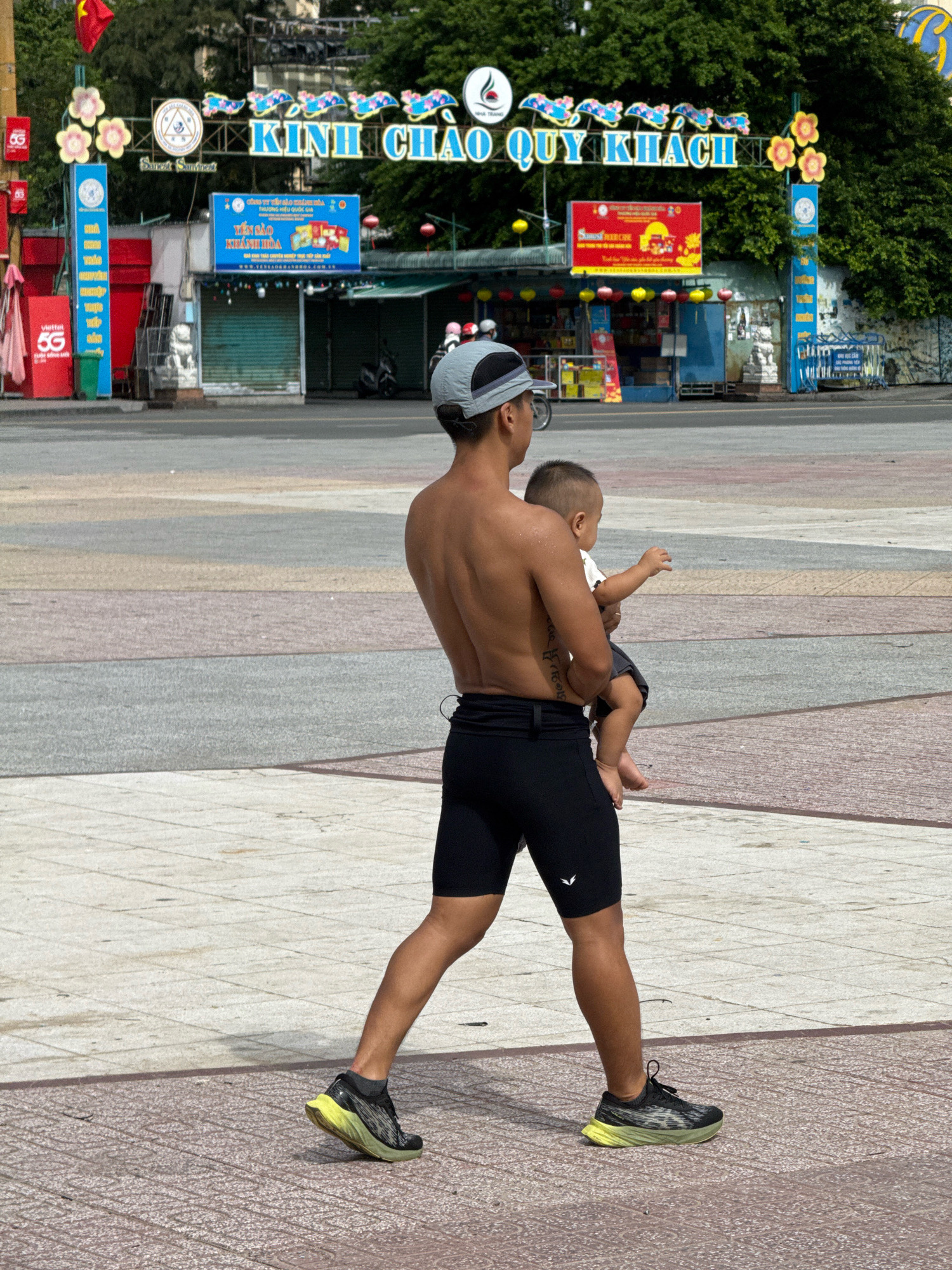 In Nha Trang, Vietnam, a shirtless man wearing a gray baseball cap and black athletic shorts walks across a paved plaza, carrying a young child in his arms. The child, wearing a white shirt and dark shorts, is looking away from the camera. The man has a tattoo on his left arm. Behind them, a large archway is adorned with blue and yellow lettering that reads "KINH CHAO QUY KHACH" which translates to "Welcome Guests". The archway is decorated with floral motifs and small lanterns. Several stalls or shops are visible behind the archway, with one displaying a red sign with yellow lettering. The plaza pavement is composed of various colored rectangular sections. The background features lush green trees and foliage.