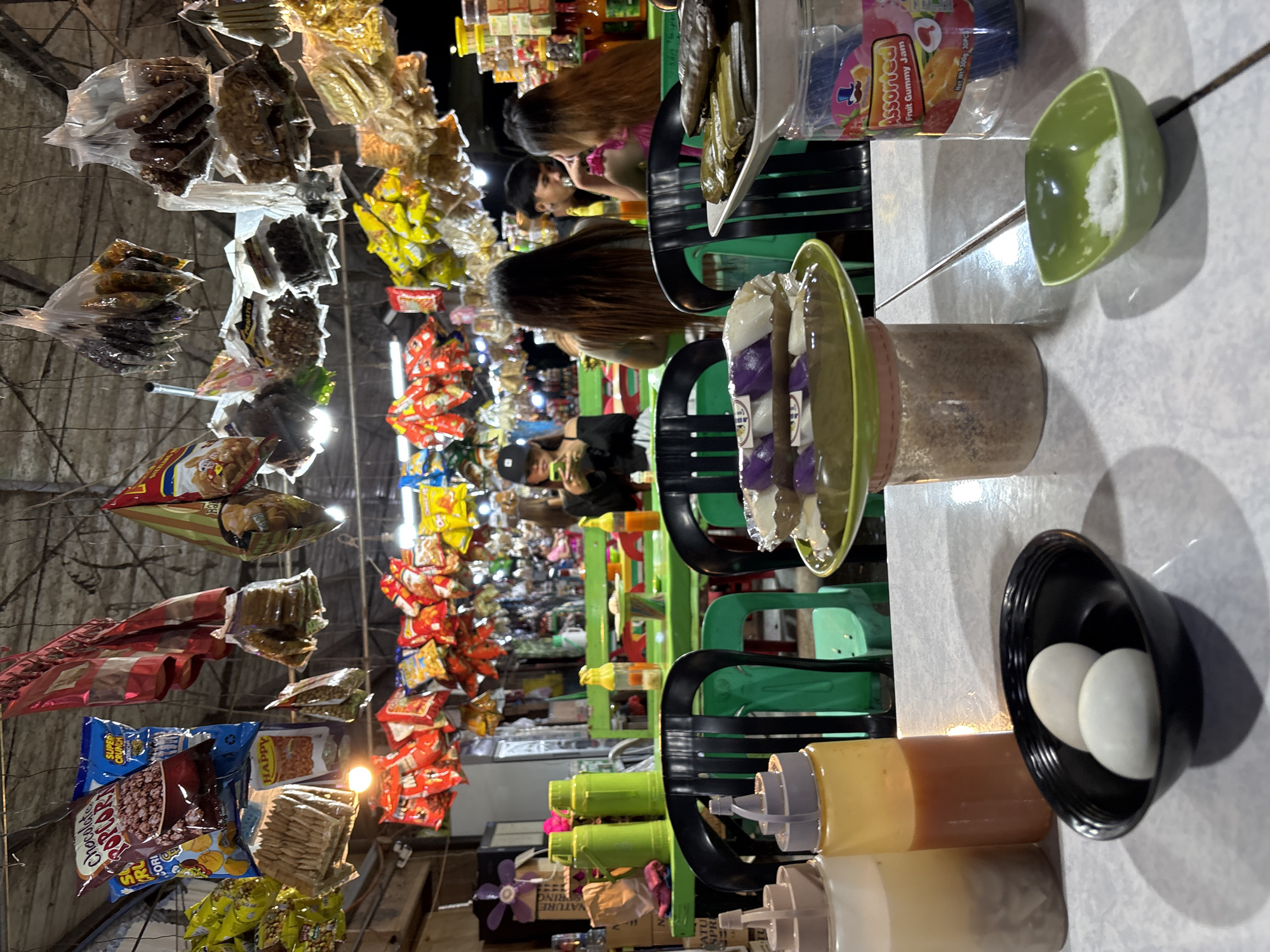 The image depicts an indoor market stall in Carmen, Philippines, with a focus on food items. Several shelves and racks are filled with an assortment of packaged snacks and goods, suspended from the ceiling. In the foreground, a table holds several dishes. A black bowl contains two white eggs. Two squeeze bottles, one clear with a light-colored liquid and another with orange sauce, are also present. A green dish rests on top of a clear plastic container, holding what appears to be rice cakes topped with purple ingredients. To the right, a plastic container labeled "Assorted Fruit Gummy Jam" is visible. In the background, several green tables and chairs are occupied by people. A woman in a black top and cap is looking at her phone. Two other individuals, a man and a woman, are seated across from each other at a table, seemingly engaged in conversation. The lighting is dim, creating an atmosphere typical of local markets.