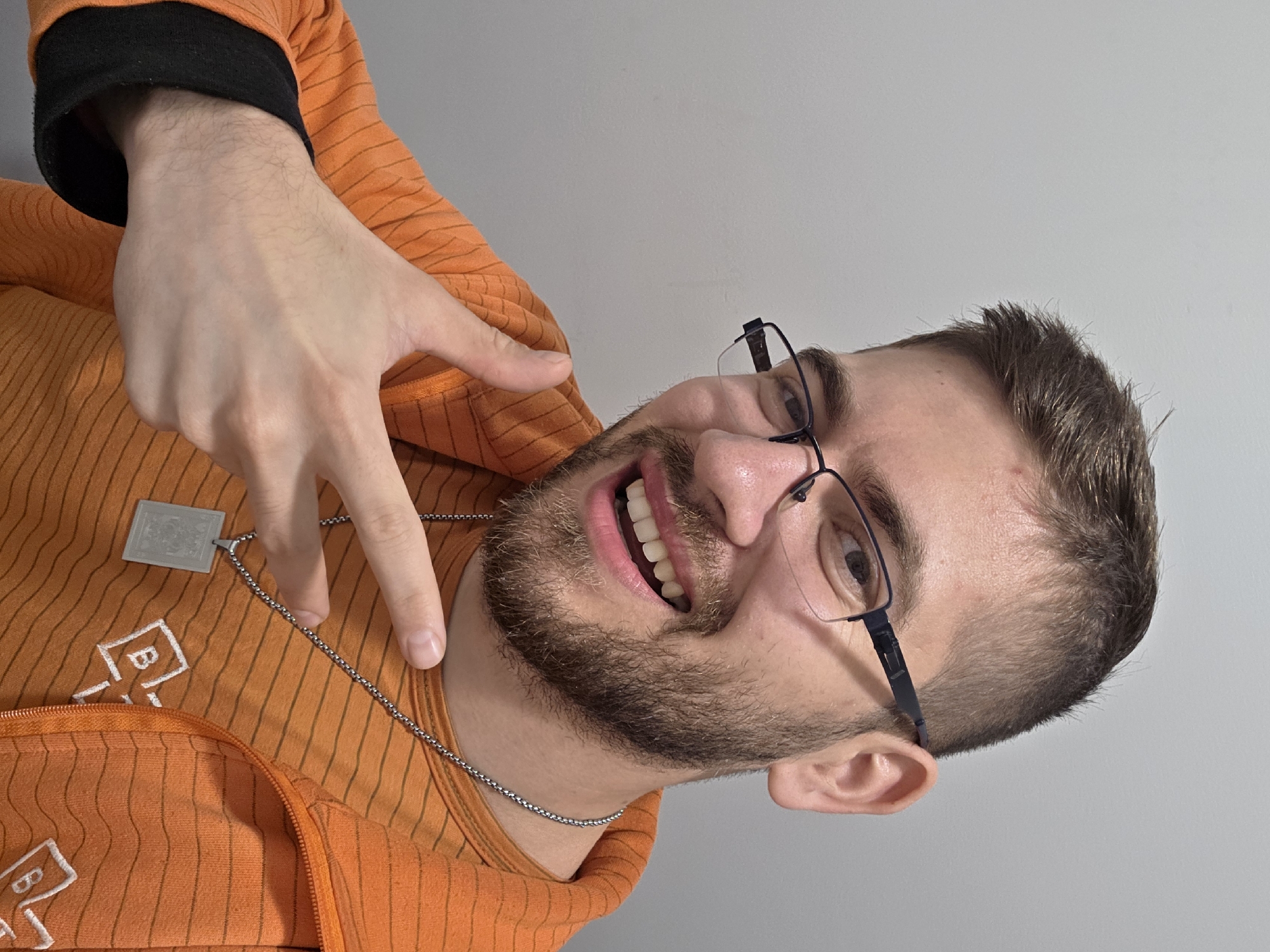 The photo is presented on a tilt, capturing a young man from an upward perspective against a plain, light gray background. He is smiling widely, looking directly at the camera with a cheerful expression. The man has short brown hair, a neatly trimmed beard, and is wearing black-framed glasses. His mouth is open, revealing his teeth.
He is dressed in an orange zip-up hoodie featuring thin dark horizontal stripes and a black cuff on his left sleeve, suggesting a long-sleeved shirt beneath. On the front of the orange hoodie, a white, stylized logo or pattern with letters resembling "B-" and another "B" further down is visible. Around his neck, he wears a silver chain necklace with a rectangular, metallic pendant. His right hand is raised, performing what appears to be a "shaka" or "hang loose" gesture with the thumb and pinky finger extended, while the middle fingers are curled.
The lighting suggests an indoor setting, and the time of day is not discernible. While the image is provided in the context of Plovdiv, Bulgaria, there are no specific visual cues within the photograph itself to indicate this location.