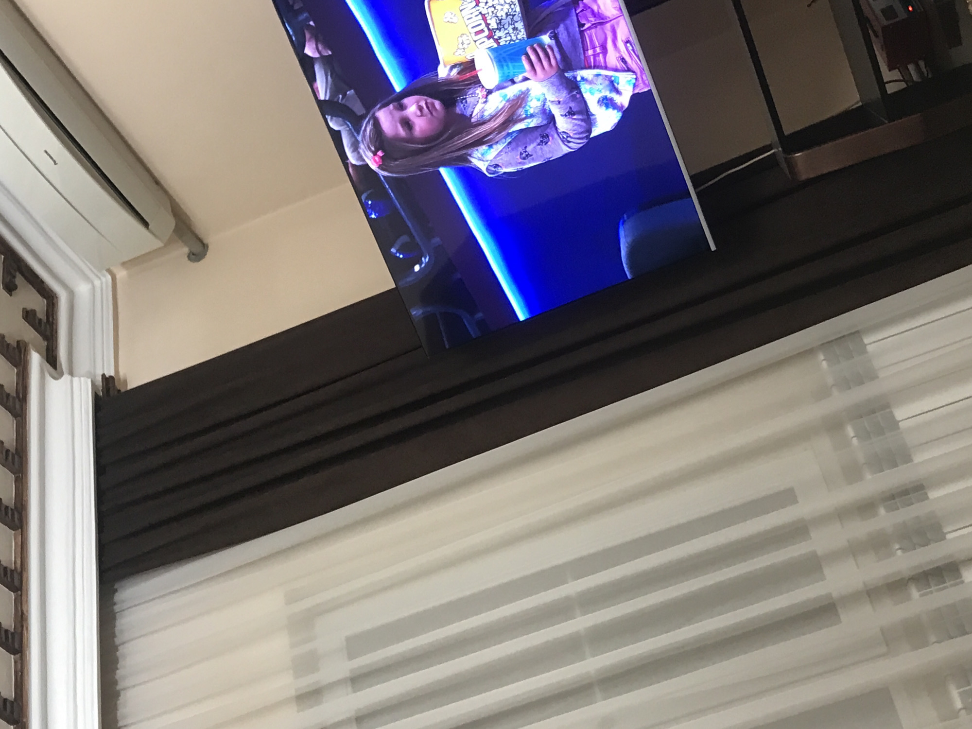 Here's a description of the image:
The image appears to be taken indoors, looking up towards a wall. A television screen is visible, displaying a scene of a young girl holding a popcorn bucket. Above the TV, there's what appears to be an air conditioning unit and a decorative molding on the wall.
Below the TV, a horizontal dark brown panel is seen above a set of white blinds or window shades covering a window.
There's no information in the image to indicate its location in Yerevan, Armenia, or any specific location.