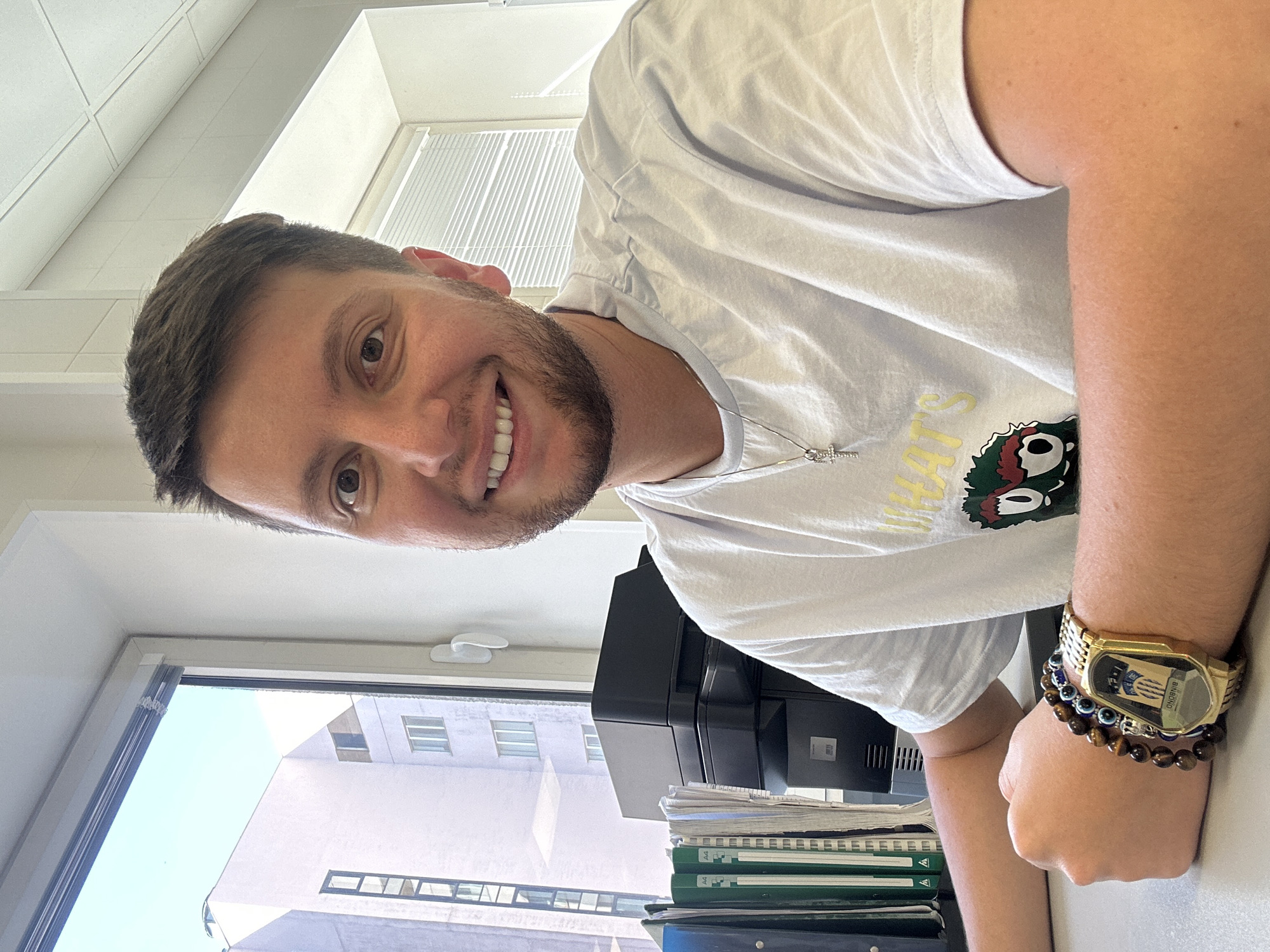 A young adult male, likely in his mid-20s to early 30s, with short dark brown hair and a light beard, is seated at a desk, smiling and looking directly at the camera. He wears a light-colored (possibly white or light grey) t-shirt featuring a partially visible green and yellow graphic that includes the word "WHAT'S" and a cartoon character. A silver chain with a small cross pendant hangs around his neck. On his left wrist, he wears a gold-toned watch with a rectangular face depicting a stylized building or emblem, along with the brand name "BINBOND." Adjacent to the watch are two beaded bracelets: one made of brown tiger's eye beads and another primarily blue and brown, including an "evil eye" bead. His forearms rest on a light grey desk surface.
To his left, on the desk, is a stack of papers and several binders (green and dark blue/black), positioned next to a black multi-function printer/scanner unit. Behind him and to his left, a window reveals an exterior view of a modern, multi-story building with a light-colored, angular facade and rectangular windows, set against a clear blue sky. The architectural style of the visible building is consistent with contemporary urban structures found within the city of Moscow, Russia. To his right and in the upper background, another window with white horizontal blinds is visible, along with white ceiling tiles in a suspended grid system. The interior walls are white.