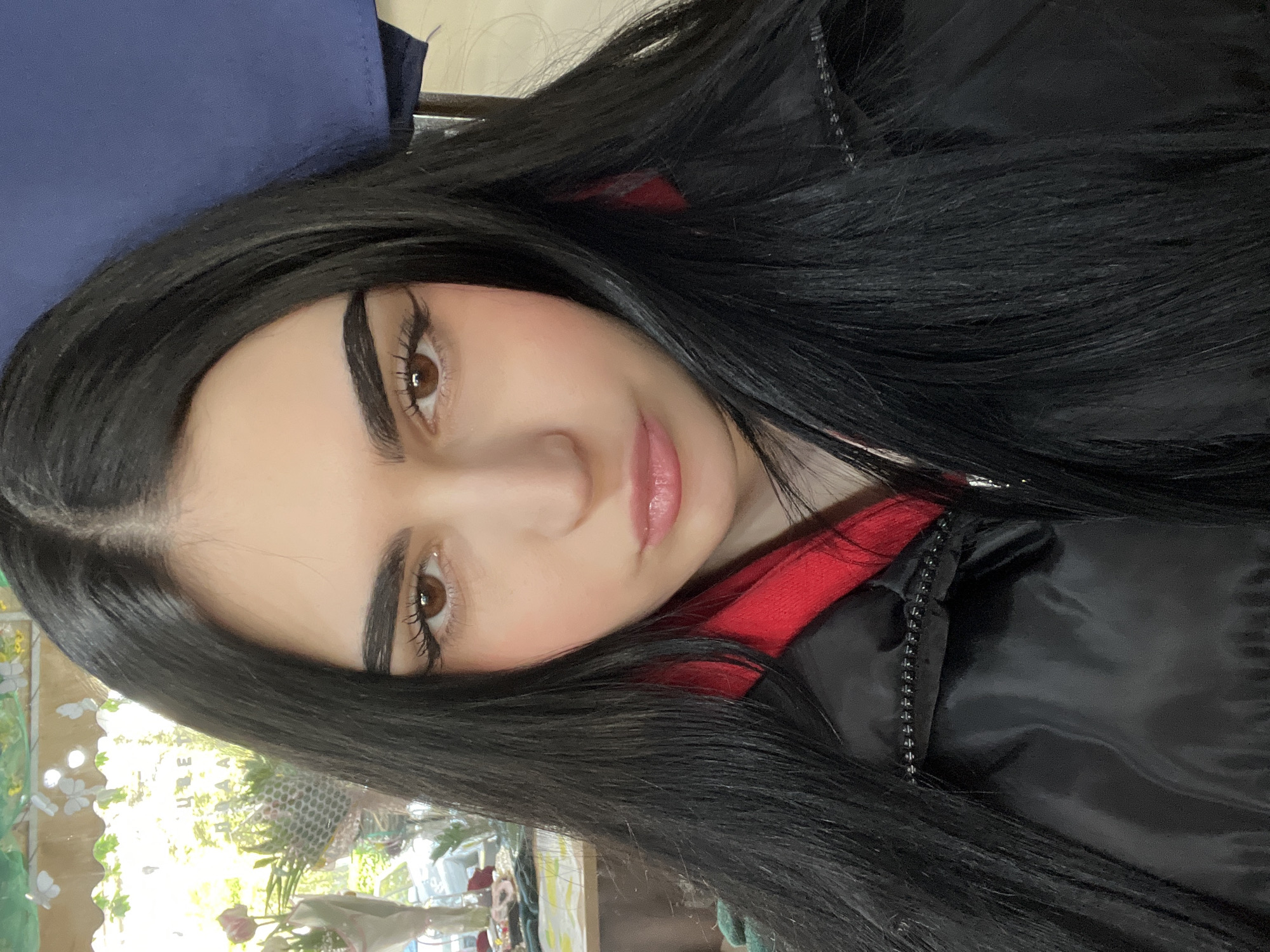A close-up, selfie-style photograph depicts a person with fair skin, light brown eyes, and black hair. The person is wearing a black jacket with a red lining visible at the collar. The background includes a blue object behind the person, and out-of-focus objects suggest an indoor environment. There is no indication of the city of Burgas, Bulgaria, in the image.