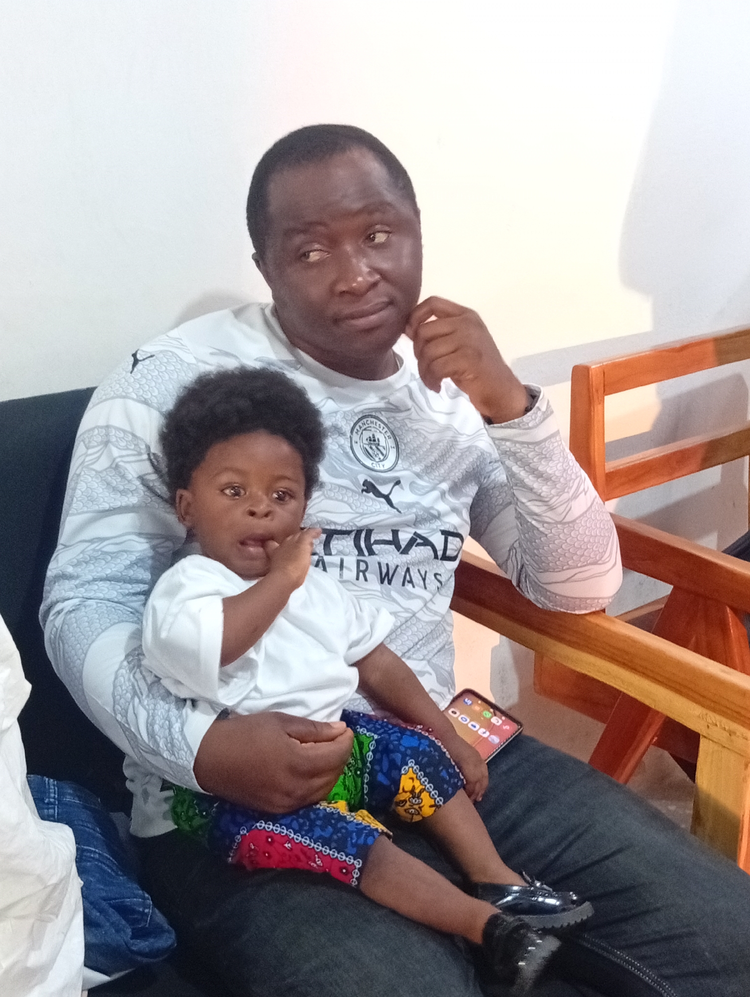 A full-shot, outdoor, eye-level, candid photograph captures a dark-skinned man holding a dark-skinned baby in his lap. The man, wearing a long-sleeved white and gray graphic t-shirt, looks to his left with his chin resting on his hand. The baby, wearing a white t-shirt and brightly colored patterned pants, looks forward with its hand near its mouth. The man is seated on a dark surface, and the baby is in his lap. To the right of the man, a wooden chair with orange-tinted slats is visible. A white wall serves as the backdrop for the scene, and a white object, possibly a bag, is seen on the left side of the frame. A cellphone is placed on the man's lap, visible between the baby's legs.