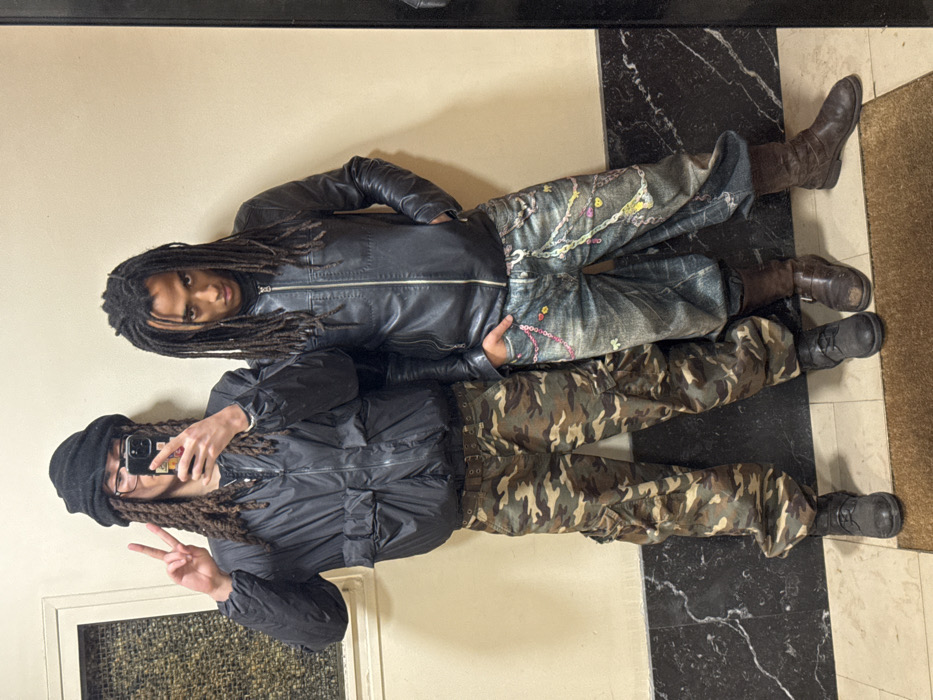 Two young males stand side-by-side indoors, posing for a mirror selfie in what appears to be a hallway or lobby in Paris, France. The male on the left, capturing the image with a dark-cased smartphone held in his right hand, wears a black puffer jacket, camouflage cargo pants, and dark shoes, with a black knit beanie covering his dreadlocks and clear-framed glasses. He forms a peace sign with his left hand. The male on the right, with long dreadlocks, wears a black leather jacket over a dark top, heavily distressed light blue jeans adorned with multiple colorful chains and small charms, and dark brown mid-calf boots. His right hand is in his jeans pocket, and he maintains a direct gaze. The background features a light cream wall with a recessed rectangular panel on the left, and a dark, polished marble floor with white veining. A dark brown doormat lies on the floor where the individuals are positioned.