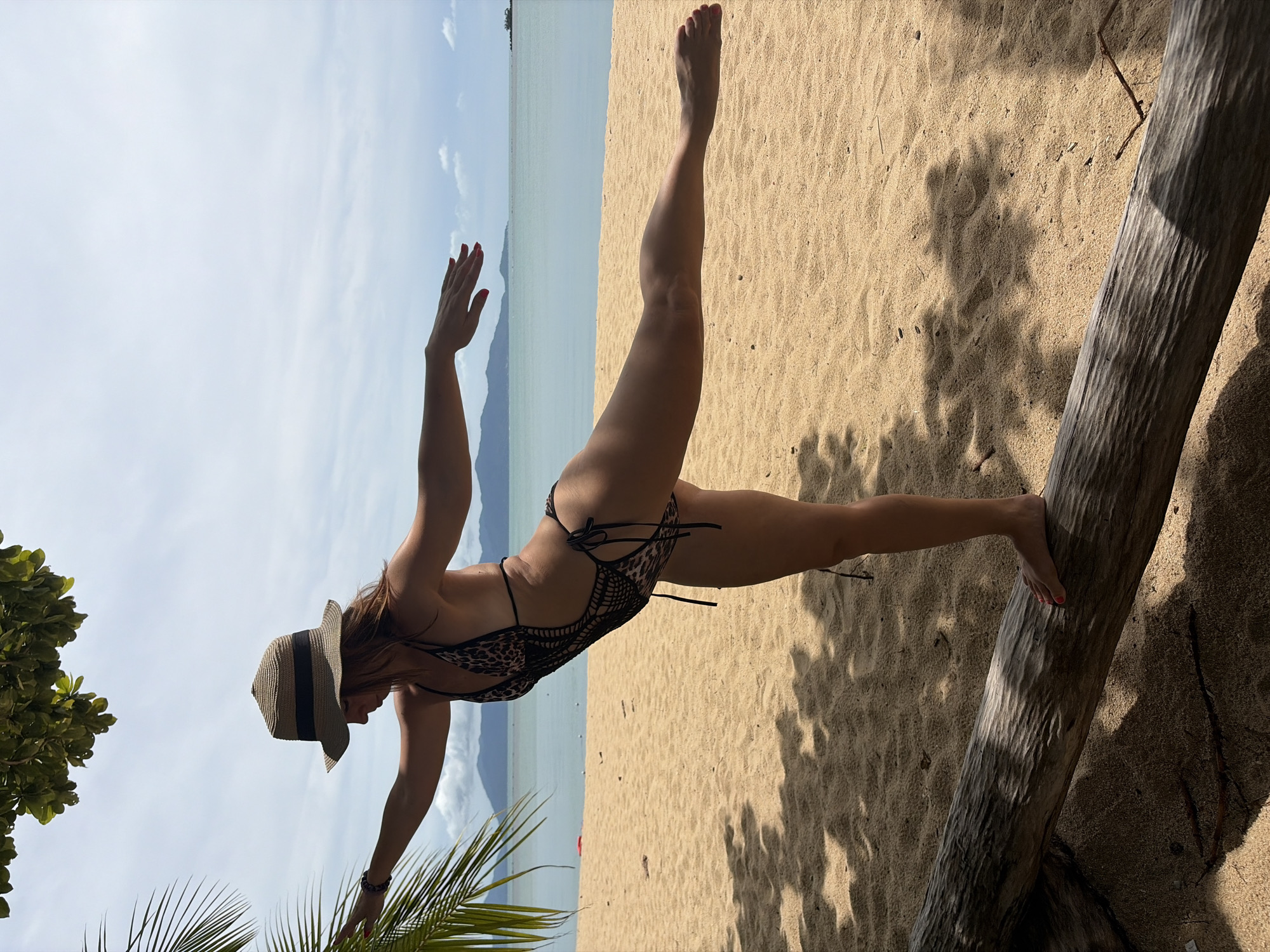 A woman stands on a fallen log on a sandy beach, balancing on one leg with her arms extended. She is wearing a leopard-print one-piece swimsuit and a woven sun hat. In the background, the turquoise ocean meets a hazy horizon with distant islands. The scene is set in Baan Bo Phut, Thailand, under a bright, partly cloudy sky. Palm fronds cast dappled shadows on the sand.