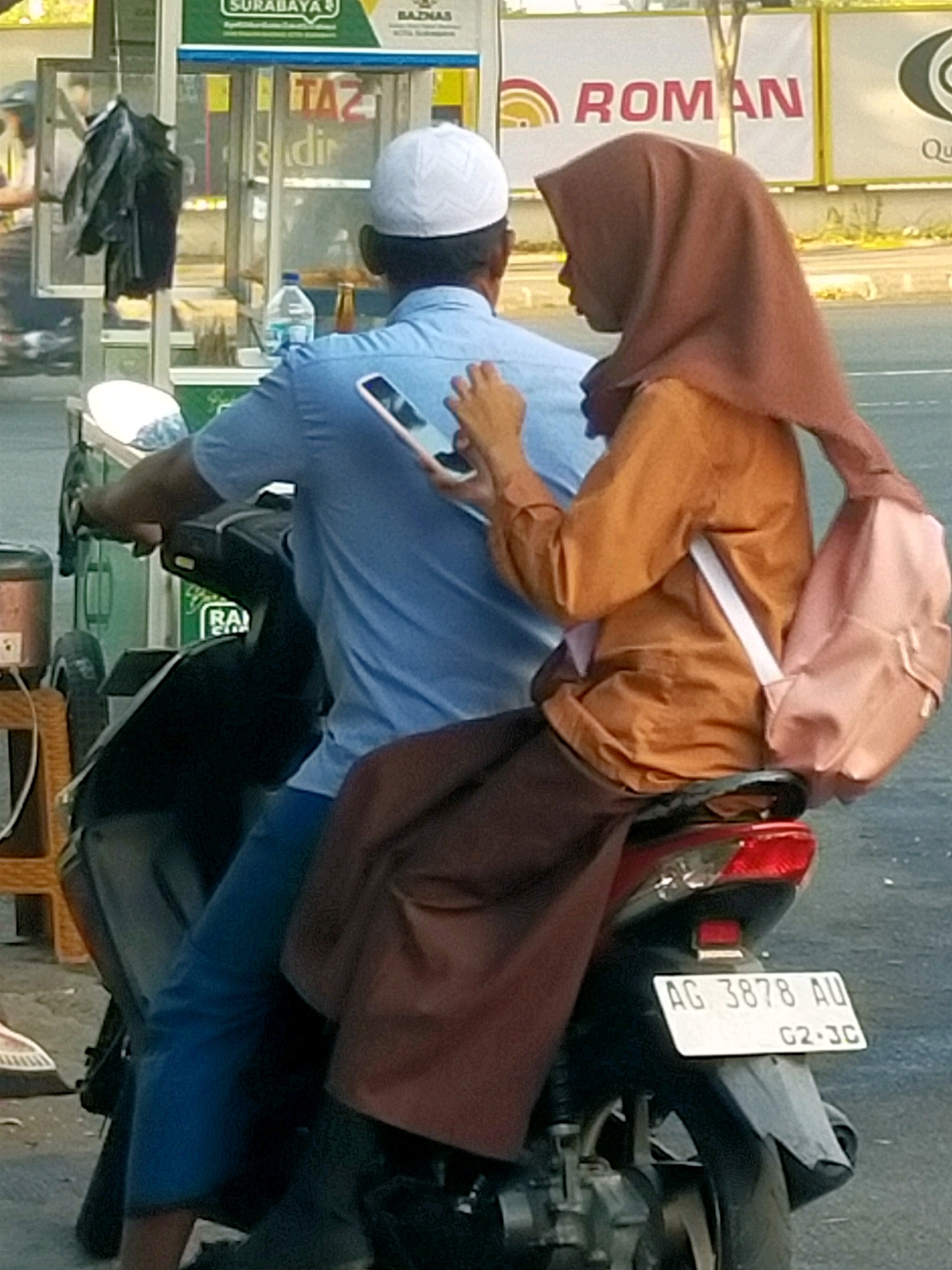 The image shows two people on a motorcycle in what appears to be a street scene in Surabaya, Indonesia. The person in the front, wearing a blue shirt and white cap, is holding a smartphone. The person in the back is wearing an orange headscarf and orange shirt and has a pink backpack. A license plate on the motorcycle reads "AG 3878 AU G2-JC". In the background, there are signs with Indonesian text, including "SURABAYA" and "ROMAN". The scene suggests a busy urban environment with commercial activity.