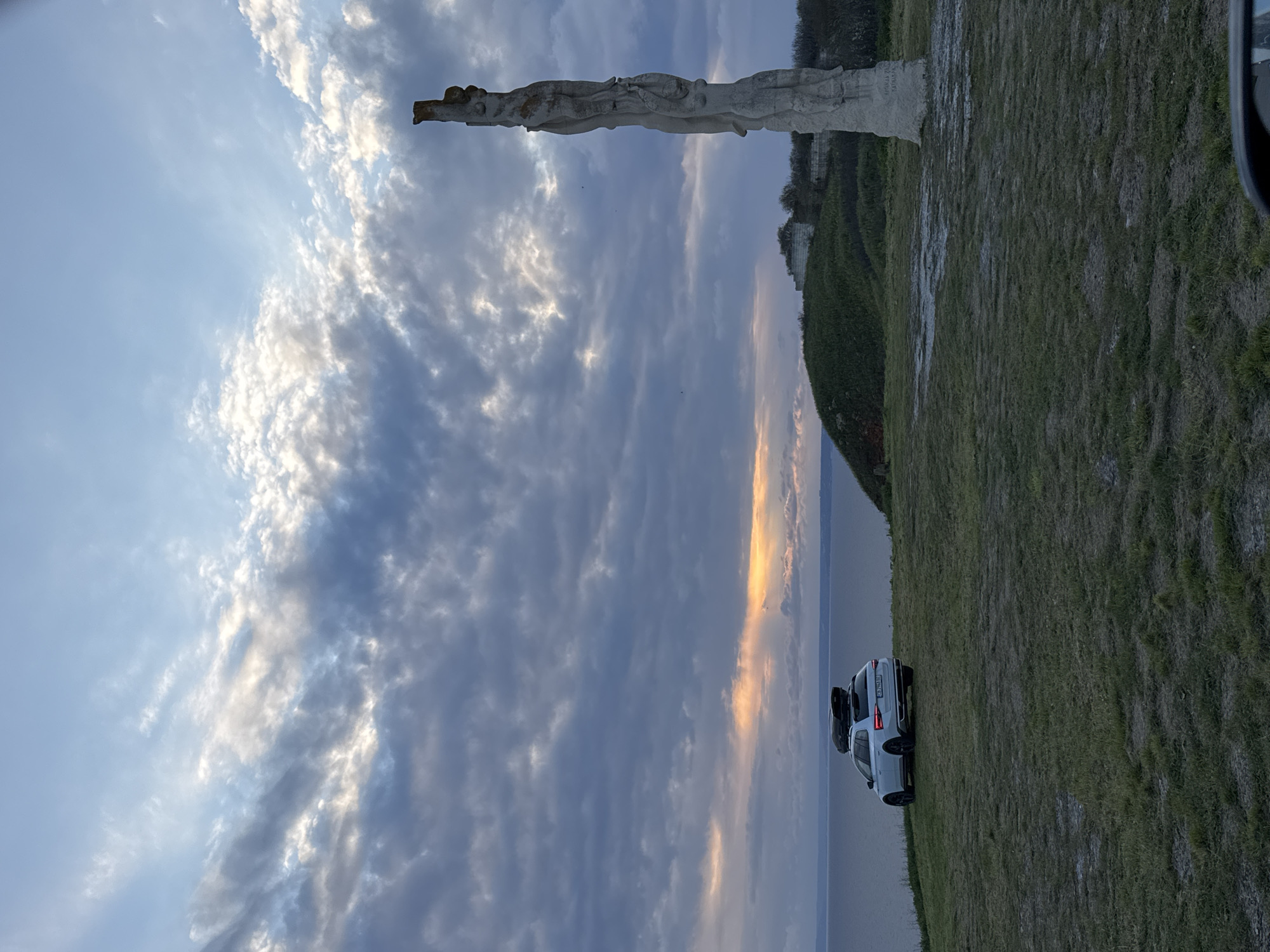 The image shows a landscape in Balgarevo, Bulgaria. In the foreground, there is a grassy field. A white car is parked near the edge, overlooking a body of water. In the background, a sculpture stands against a partly cloudy sky. A golden hue is present in the sky above the water.