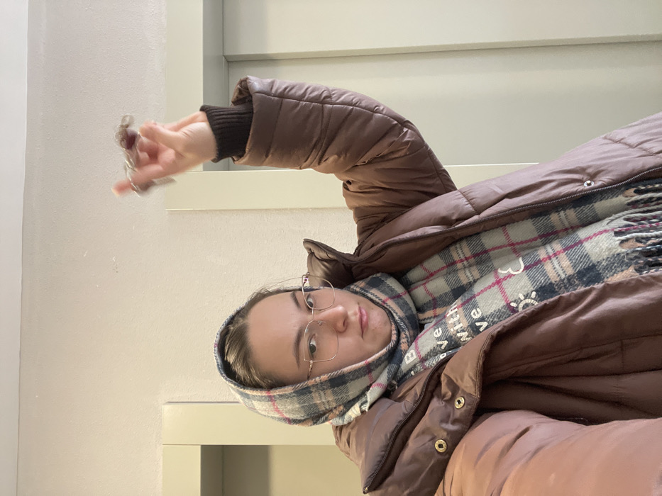 An individual is depicted indoors in Kupavna, Russia, standing in what appears to be a hallway or common area of an apartment building, indicated by two light-colored vertical door frames or elevator panels on either side of the background. The walls and ceiling are plain and light-colored. The subject is a female wearing eyeglasses, a brown puffer jacket, and a plaid scarf draped over her head and neck. The scarf features a pattern of light blue/grey, dark blue, pink, and white, with text "BELIEVE WITH LOVE" and small heart and sun icons. Her right arm is raised, bent at the elbow, and her hand holds a set of keys with multiple keyrings, one appearing dark and textured, the other metallic. The keys are slightly blurred due to motion. The subject maintains a neutral to slightly stern facial expression.