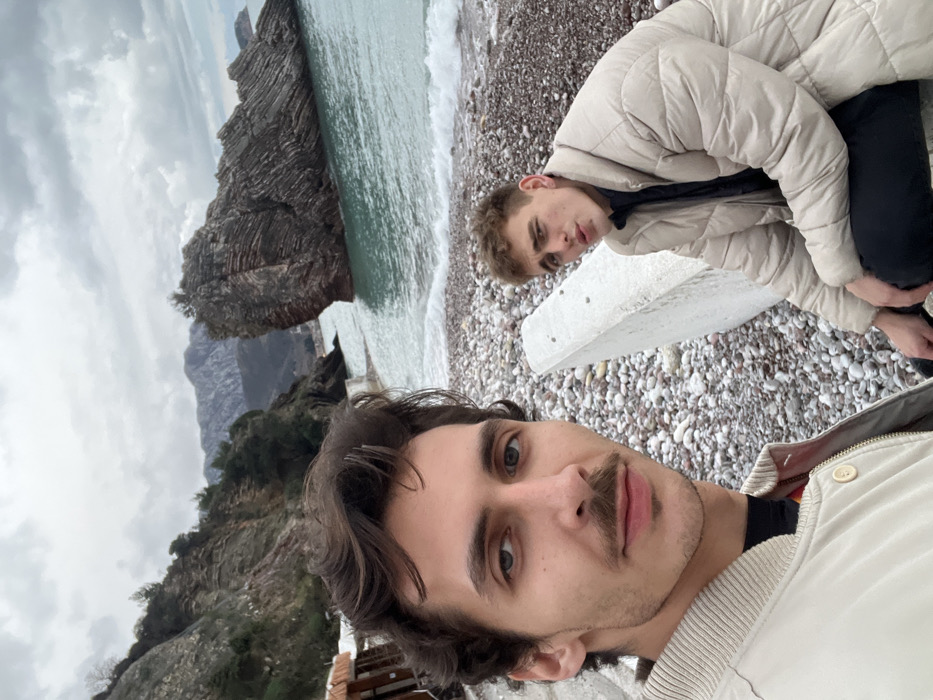 The image is a selfie-style photograph taken outdoors, rotated 90 degrees clockwise. It features two young men on a pebble beach with a rocky coastline and the sea in the background under a cloudy sky.
In the immediate foreground, facing directly towards the camera, is a young man with dark, wavy hair and a noticeable mustache. He is wearing a light-colored jacket or sweater over what appears to be a dark t-shirt. His expression is neutral, and his eyes are looking directly into the lens.
Slightly behind and to the left of the foreground man, closer to the water, is another young man. He has lighter hair and is wearing a puffy, light beige or off-white jacket with dark pants. He is in a semi-crouching or leaning position, looking towards the camera with a neutral expression.
Between the two men, on the pebble beach, a large, light-colored concrete block or barrier is visible. The beach itself is covered with numerous small, light-colored pebbles.
To the left side of the image, the main features of the landscape are visible. A significant dark, layered rock formation or cliff juts out into the greenish-blue sea. Beyond this rock, more mountainous terrain or cliffs extend into the distance under the overcast sky. Small waves are breaking gently on the shore. The sky is predominantly cloudy, indicating an overcast day.