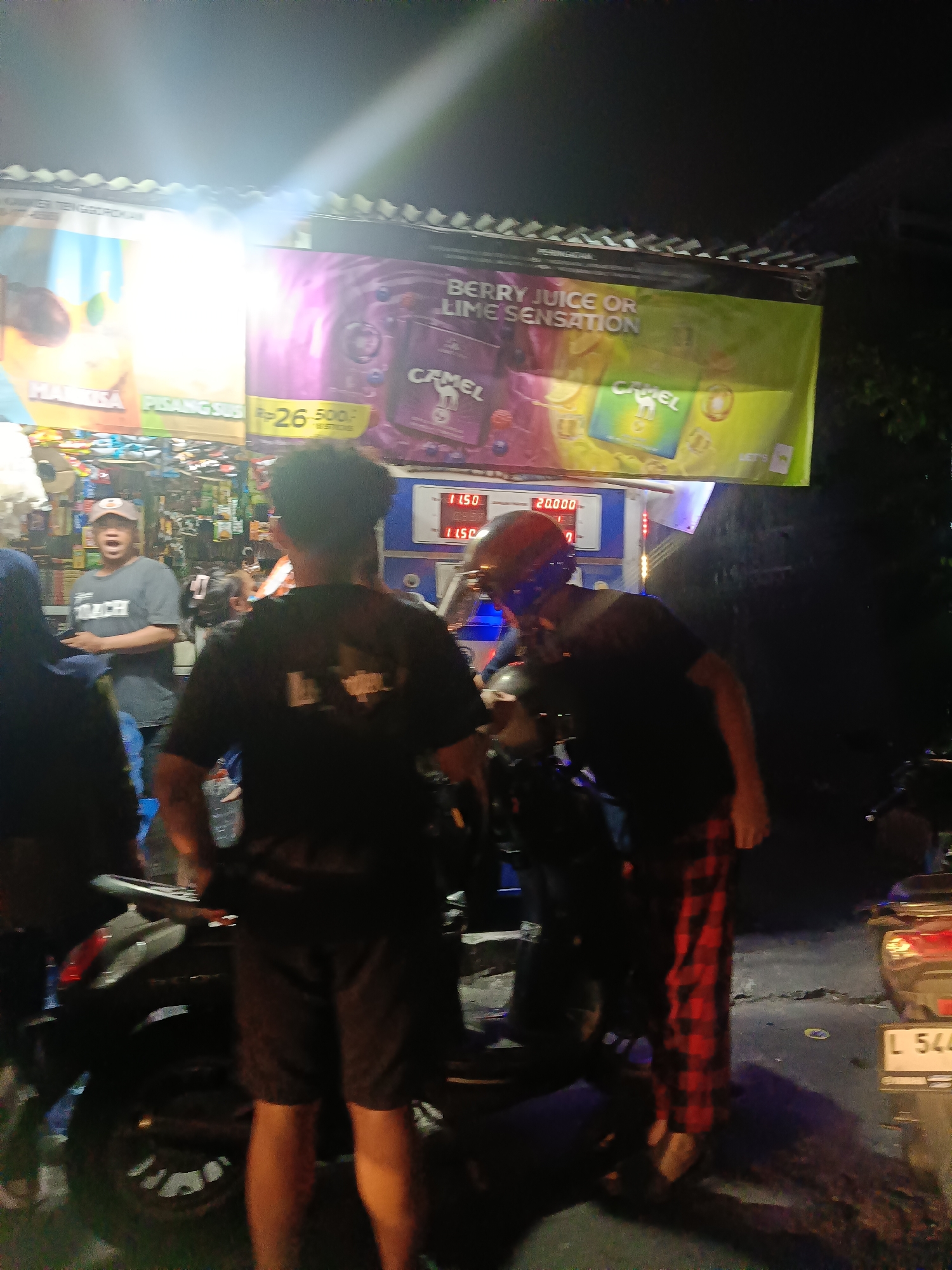 The image depicts a nighttime scene outside a store in Surabaya, Indonesia. A brightly lit banner advertising "BERRY JUICE OR LIME SENSATION" with "CAMEL" branding is prominent. In the foreground, two individuals are near motorcycles. One person, wearing a black t-shirt and plaid pants, is leaning towards a motorcycle. Another person, partially visible, appears to be wearing a black t-shirt. Further back, another individual in a grey t-shirt with the word "COACH" and a baseball cap is visible, seemingly interacting with someone in the store. The store appears to be a small retail establishment with various products displayed inside. The ground is asphalt, and there is a license plate visible on a motorcycle in the right portion of the image.