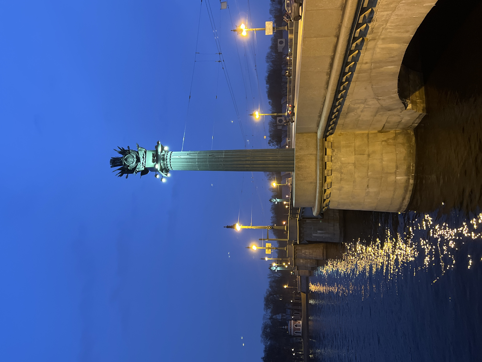The image depicts the Birzhevoy Bridge over the Malaya Neva River in Saint Petersburg, Russia, during twilight or early night. The sky is a deep, clear blue, transitioning to a slightly lighter shade near the horizon.
In the foreground, on the right side of the frame, stands a prominent, teal-colored decorative lamppost column. This column features a fluted shaft, a base, and a capital adorned with a circular medallion containing a profile bust, surrounded by a radial, wing-like motif, and stylized elements resembling ship prows (rostra). Small white lights are affixed to the capital, illuminating the upper portion of the column.
The multi-arched stone bridge, constructed of light-colored blocks, extends across the scene from the right foreground to the left mid-ground. Its roadway is lined with numerous regularly spaced streetlights emitting a warm yellow-orange light. The light from these streetlights reflects off the dark surface of the Malaya Neva River in the lower-left, creating elongated, shimmering patterns.
Vehicular traffic, including several cars with visible headlights and taillights, moves across the bridge. Indistinct pedestrian figures are visible on the right side of the bridge's walkway. A network of dark overhead electrical wires is visible traversing the sky above the bridge.
On the distant left bank, a line of bare trees is visible, behind which stands a distinct, white-walled building with a prominent red roof, resembling a historical pavilion or a part of the Old Saint Petersburg Stock Exchange complex (now the Central Naval Museum). The overall scene is characterized by artificial illumination contrasting with the ambient twilight.