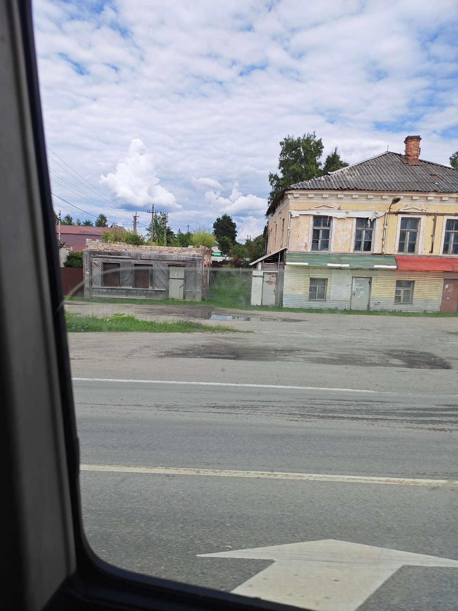 The image depicts a street scene in Едрово, Russia, on a cloudy day. The perspective is from inside a vehicle, looking out through the window, which frames the right side of the scene.
The main subjects are two buildings. On the left, there is a small, dilapidated wooden structure with a weathered appearance, featuring two boarded-up windows and a door. It appears to be abandoned or in disrepair. To the right of this, and slightly further back, stands a larger, two-story building. The upper floor is painted a pale yellow, with multiple windows and decorative architectural details. The lower floor is clad in horizontal planks painted in sections of green, yellow, and blue, with a red awning-like element on the right. This building also appears aged, with peeling paint and some visible damage. A brick chimney rises from the roof of this building.
In front of the buildings is a paved area, likely a road or a parking lot, which appears wet in places, suggesting recent rain. The road has tire marks and a faded white line. A large white arrow, pointing to the right, is painted on the road in the foreground, indicating a directional lane. Beyond the buildings, trees and power lines are visible, with a cloudy sky overhead. The overall impression is of a rural or small-town setting, possibly in a state of gradual decline or undergoing transition, given the contrasting conditions of the two buildings. The weather is overcast, but there are patches of blue sky and distinct white clouds, suggesting a dynamic weather pattern. No people are visible in the frame. There is no visible text.
