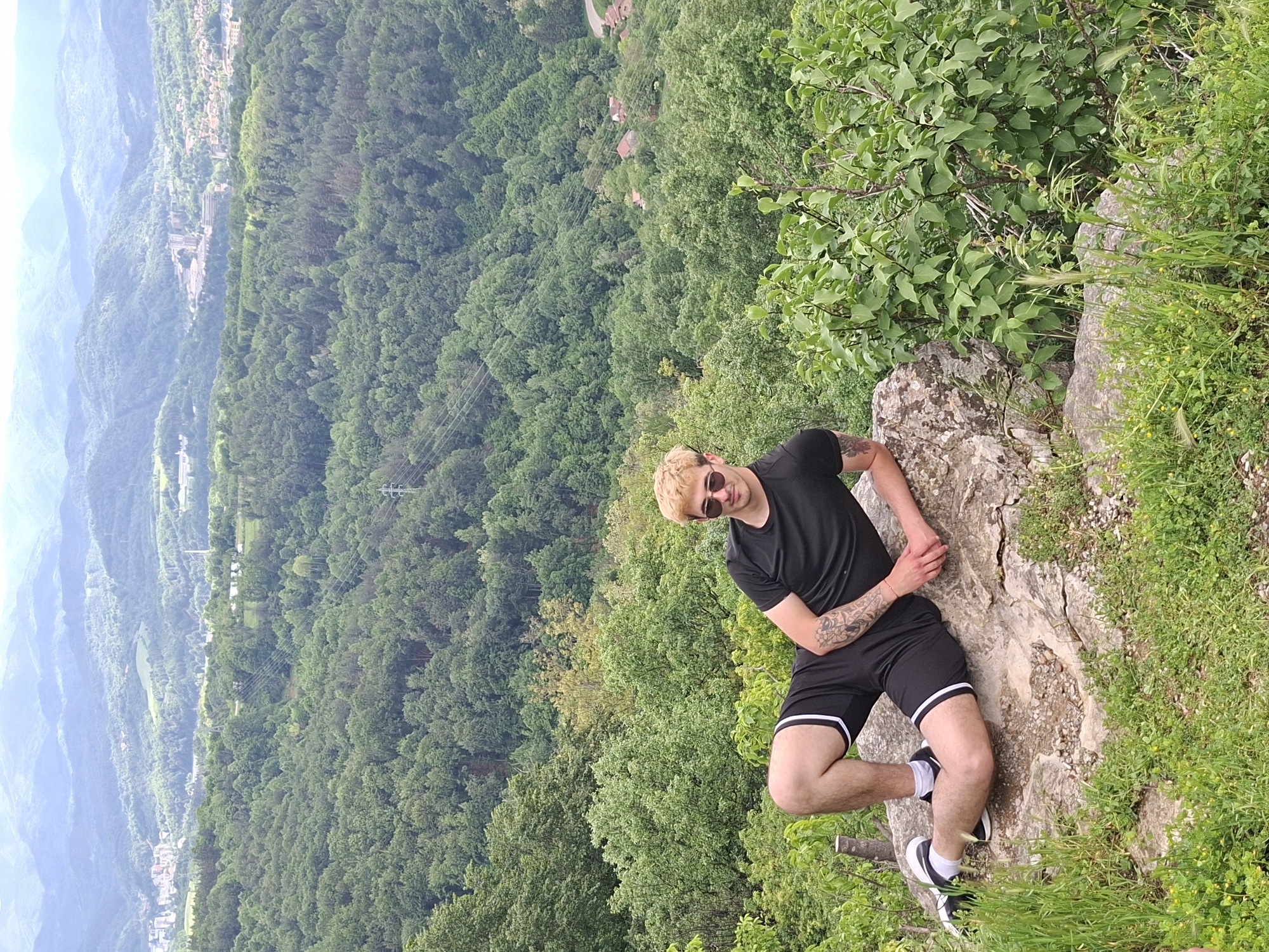 The image shows a young man with blond hair, wearing sunglasses, a black t-shirt, and black shorts with white trim. He is sitting on a rock at a high vantage point, overlooking a densely forested valley. The foreground includes foliage and grassy areas, indicating a natural setting. In the distance, through a break in the trees, are mountains and a portion of what appears to be a city or town. Based on the location information, the scene is likely to be in or near Gabrovo, Bulgaria. The man is posing for the camera, looking directly at the viewer with a slight smile.