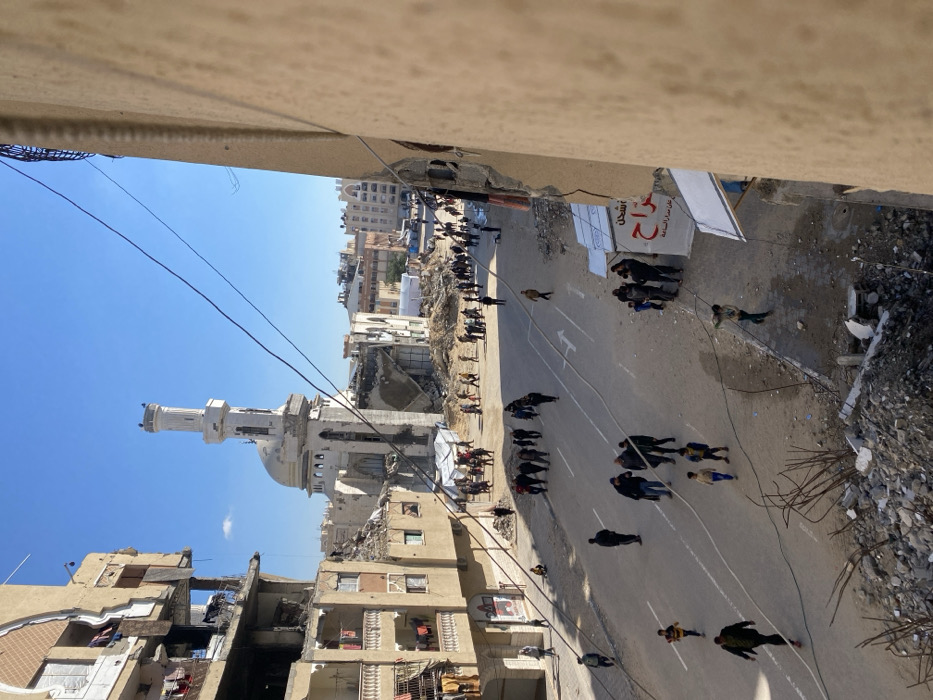 The image captures a wide street scene in the city of Khan Yunis, Palestinian Territory, displaying widespread structural damage under a clear blue sky. In the mid-ground, the Great Mosque of Khan Yunis is prominently visible, exhibiting significant damage; its minaret shows fractures and missing sections, while the main mosque structure has suffered partial collapse, exposing internal elements. Buildings lining both sides of the street are extensively damaged; the multi-story residential structure on the left has large sections of its facade collapsed, revealing interior rooms and balconies, some with clothing hanging. The building partially seen in the immediate foreground on the right also shows damage, with debris accumulated at its base.
The asphalt street, marked with white directional arrows, is populated by numerous individuals. Predominantly adults, along with several children, are observed walking in various directions across the road. A young child in a yellow shirt is running in the lower-left quadrant. Along the edges of the street, piles of rubble and debris are present. Further down the street, amidst more damaged buildings, several white tent-like structures are visible. Overhead, multiple utility cables crisscross the upper portion of the frame.