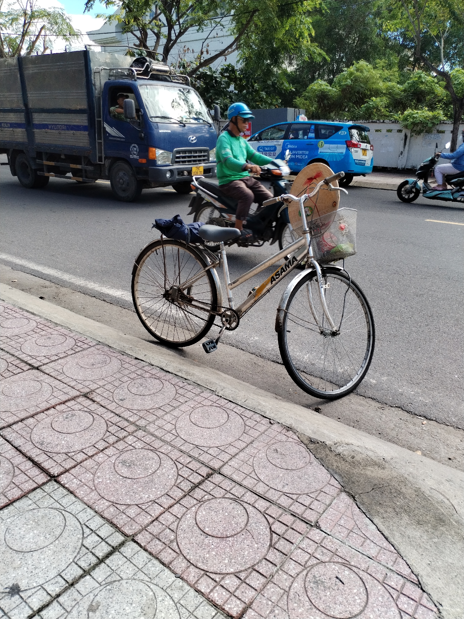 This image captures a vibrant street scene in Nam Nha Trang Ward, Vietnam, depicting a sunny daytime setting. The main subject is a silver "ASAMA" bicycle leaning against a concrete curb in the foreground, between a decorative tiled sidewalk and the asphalt road. A straw conical hat rests on its handlebars, and a wire basket on the front contains some visible items. A blue fabric item is draped over the rear fender.
In the mid-ground, a blue "HYUNDAI" truck with a large cargo bed is moving from left to right, its driver partially visible. The truck's license plate reads "79C 028.30", and the word "HÀNG" is partly visible on its side. To the right of the truck, a person in a green long-sleeved shirt and blue helmet rides a motorcycle. Further right, a blue "Midea" branded taxi/van, also displaying "TAXI QT", "INVERTER ON MIDEA", and the numbers "52 52 52", is driving. The number "813" is also visible on its rear. On the far right, another person on a scooter is partially visible.
The scene is framed by lush green trees and glimpses of white buildings in the background. The sidewalk in the foreground features light pink and grey square tiles, each decorated with concentric circular patterns. The image conveys a sense of everyday urban activity and traffic.