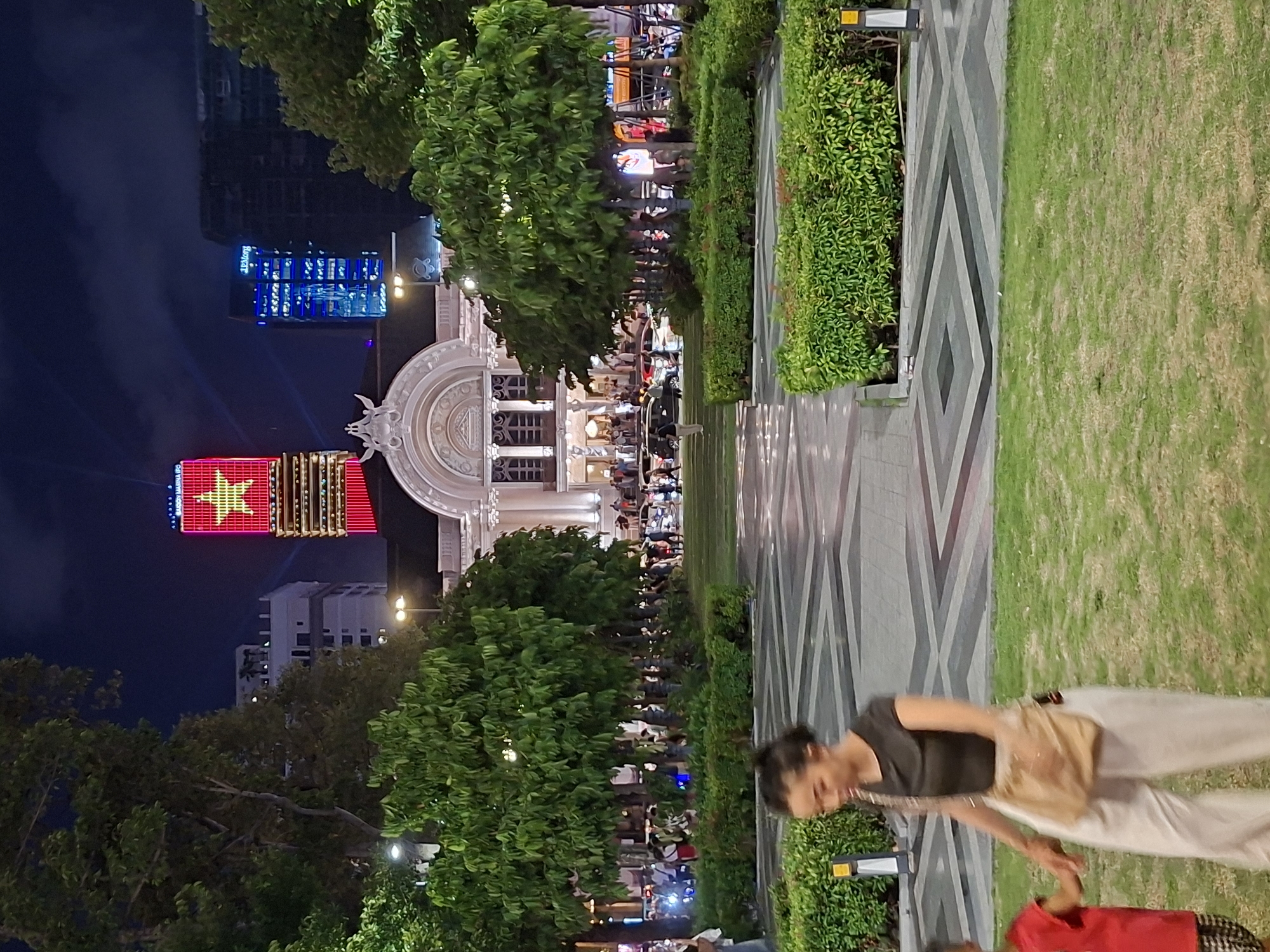 A nighttime scene captures the Ho Chi Minh City Hall in Thủ Đức, Vietnam, illuminated by external lights. A prominent skyscraper in the background is adorned with a large red Vietnamese flag with a yellow star. The foreground features a woman and a child walking on a grassy area in front of patterned pavement and manicured hedges. Several trees are visible, and a crowd of people and vehicles are present in the middle ground, suggesting a public space or event. The sky above is dark with scattered clouds.