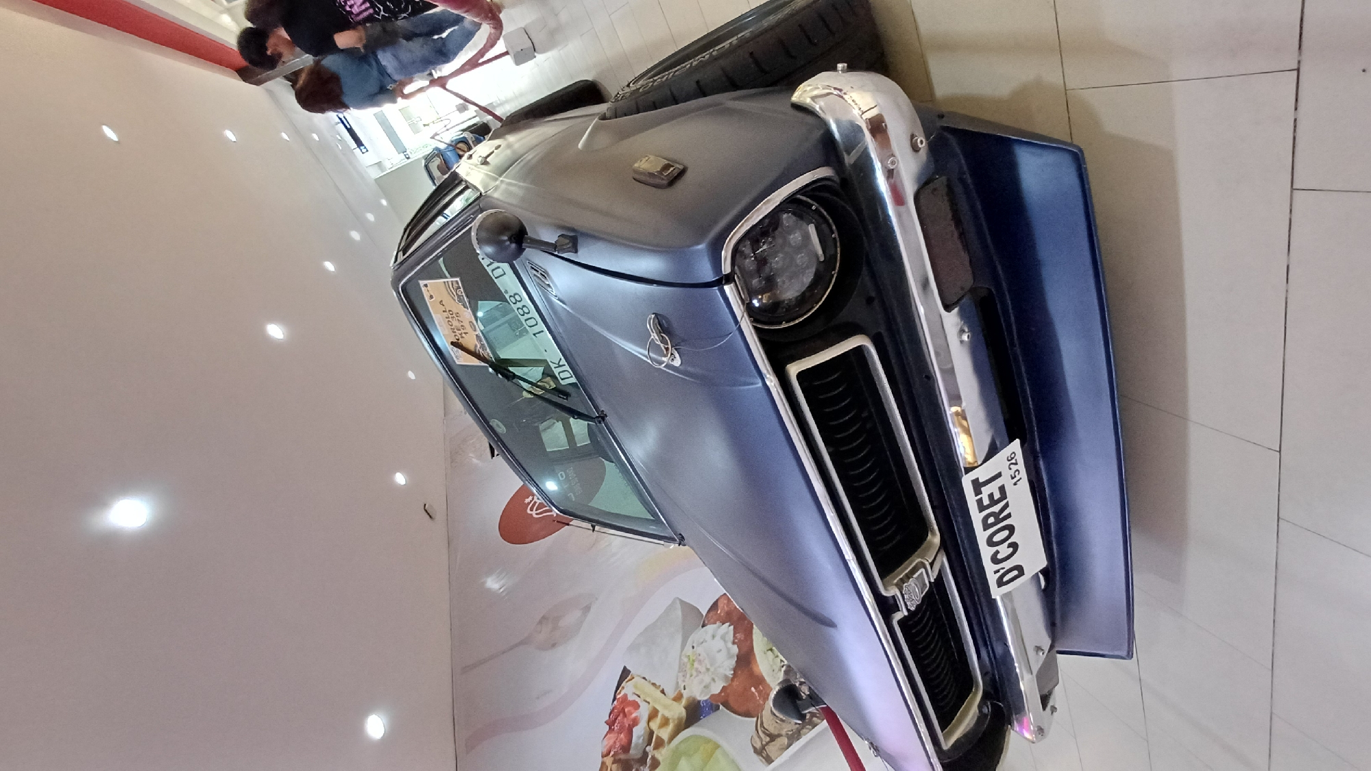 The image shows a blue vintage car, possibly a modified Toyota Corolla 1970, on display indoors. The car has a license plate that reads "D'CORET 1526." A sticker on the windshield identifies the car as a "COROLLA 1970," accompanied by Indonesian text. The car is positioned in front of a wall with a food-themed design, possibly indicating a restaurant or food-related establishment. In the background, people are visible, indicating the location may be a public space such as a mall. The text "Kuta, Indonesia" is not explicitly visible in the image.