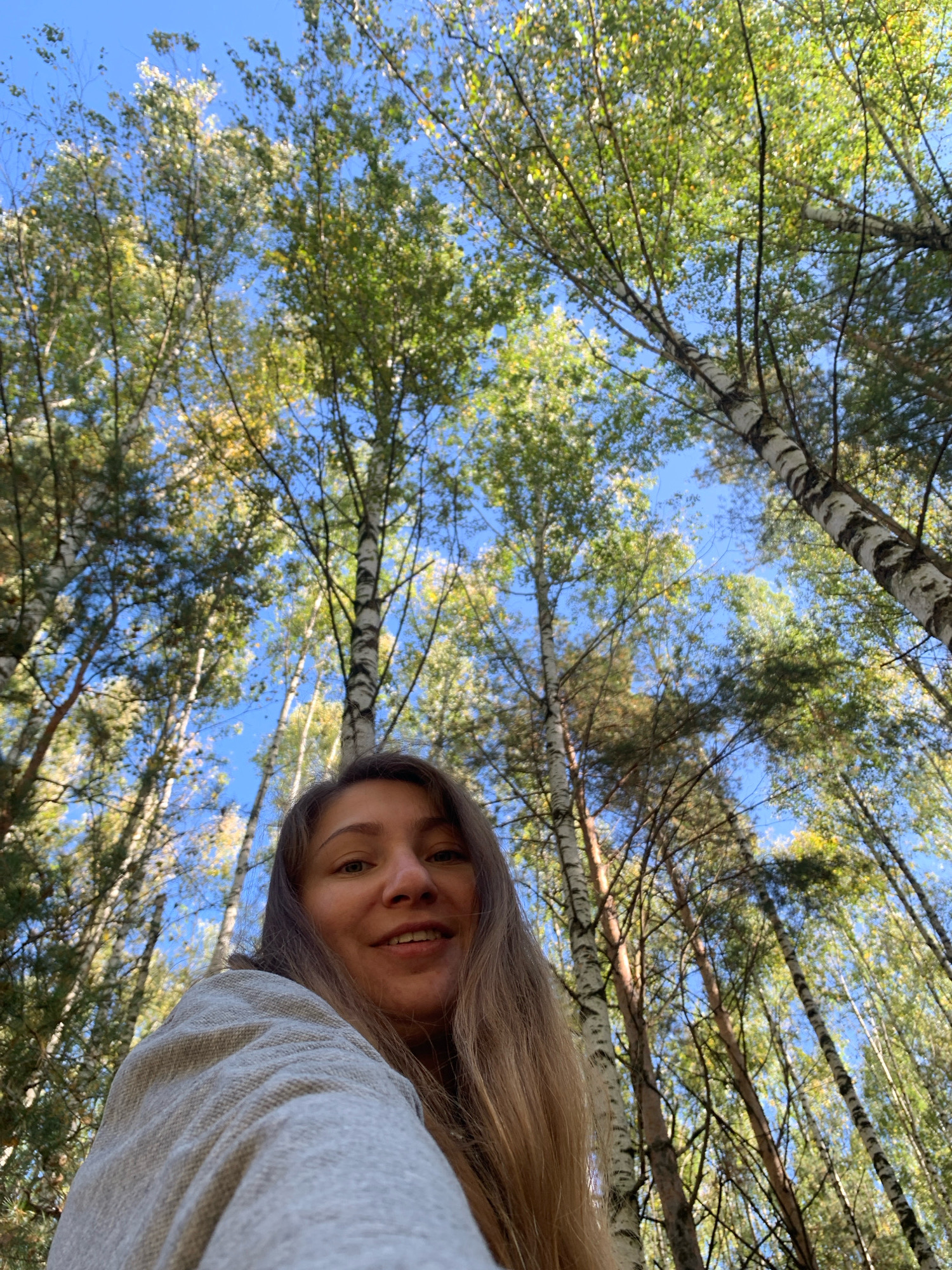 The image is a low-angle selfie taken outdoors in a wooded area, likely a park or forest, under a clear blue sky. The foreground features a woman with long, light brown hair, wearing a textured grey garment. She is smiling, looking towards the camera. Behind her, the scene is dominated by tall trees with white and dark bark, characteristic of birch trees, and lush green and yellow foliage, suggesting it is autumn. The perspective emphasizes the height of the trees and the open sky. There is no indication of this being specifically in the city of Chaikovsky, Russia.