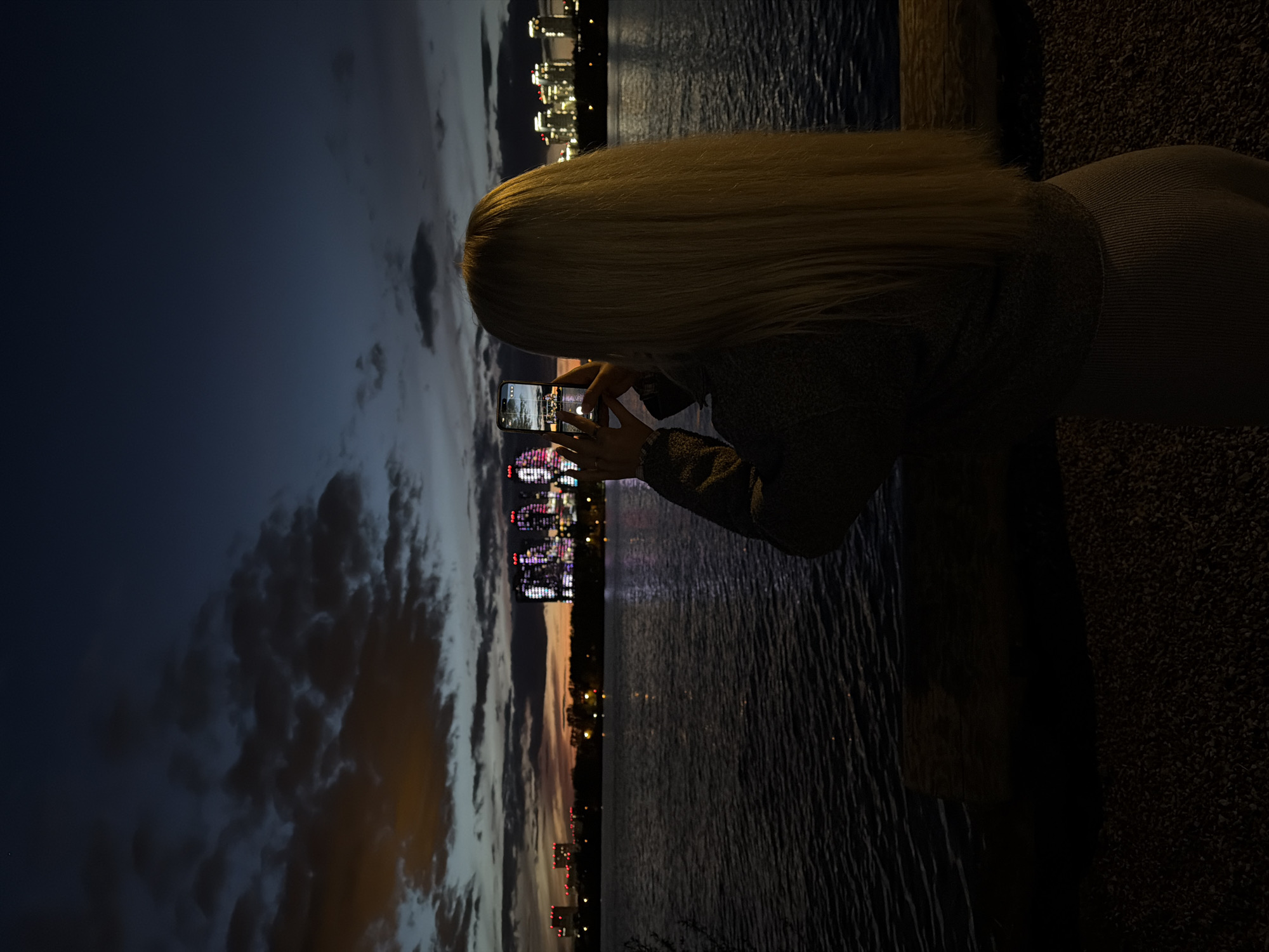 A young woman with long, blonde hair stands near a body of water at night, taking a photo with her phone. The photo appears to be of the Kazan Family Center building in Kazan, Russia, illuminated with colorful lights. The sky is dark with clouds, and city lights are visible in the background across the water. The woman is wearing a dark, textured top and pants, and she is positioned to the right of the frame.