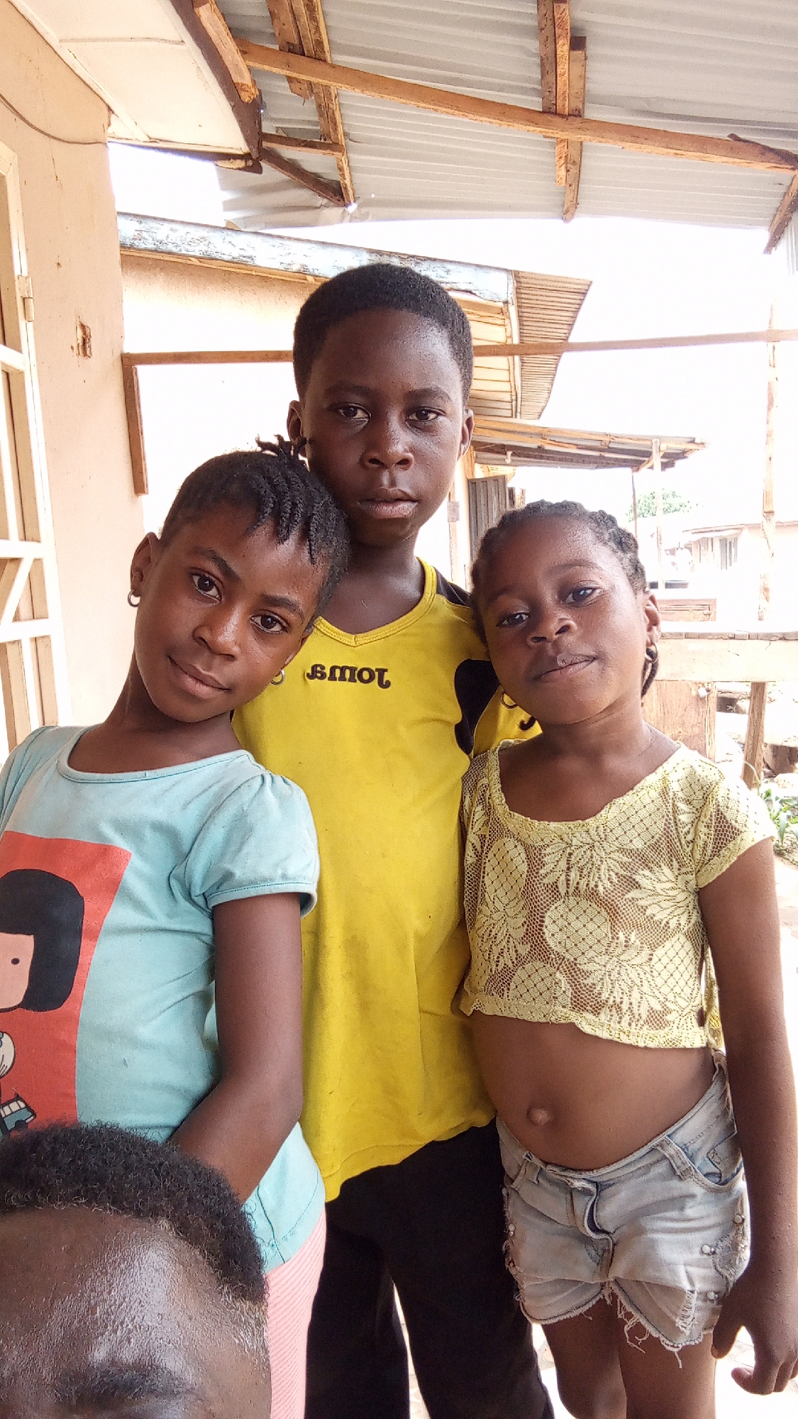 Three children are standing in front of a partially-constructed building in Sabon Tasha, Nigeria. The child in the center wears a yellow shirt with black accents. To the left, a child wears a light blue shirt with a cartoon design. To the right, a child wears a yellow lace crop top and denim shorts. A portion of an adult head is visible in the lower left corner of the image. Overhead, a corrugated metal roof is supported by wooden beams.