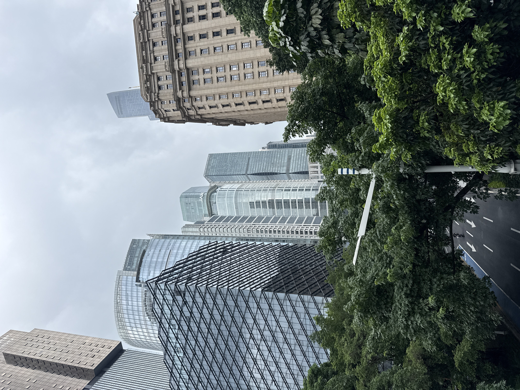 The image captures a view of modern and traditional architecture in Tianhe District, China, under an overcast sky. In the foreground, lush green trees partially obscure a street with white lane markings and directional arrows. A single motorcycle is visible on the road. Behind the trees, a cluster of skyscrapers dominates the skyline. These include a building with a distinctive geometric glass facade, a curved glass skyscraper, and several other contemporary high-rises. To the right, a multi-story building with a classical architectural style, featuring numerous windows and ornate detailing, stands prominently. Another sleek, tall glass tower is seen rising behind it. The overall scene suggests a dynamic urban environment where contemporary development coexists with older structures, all under a muted sky.