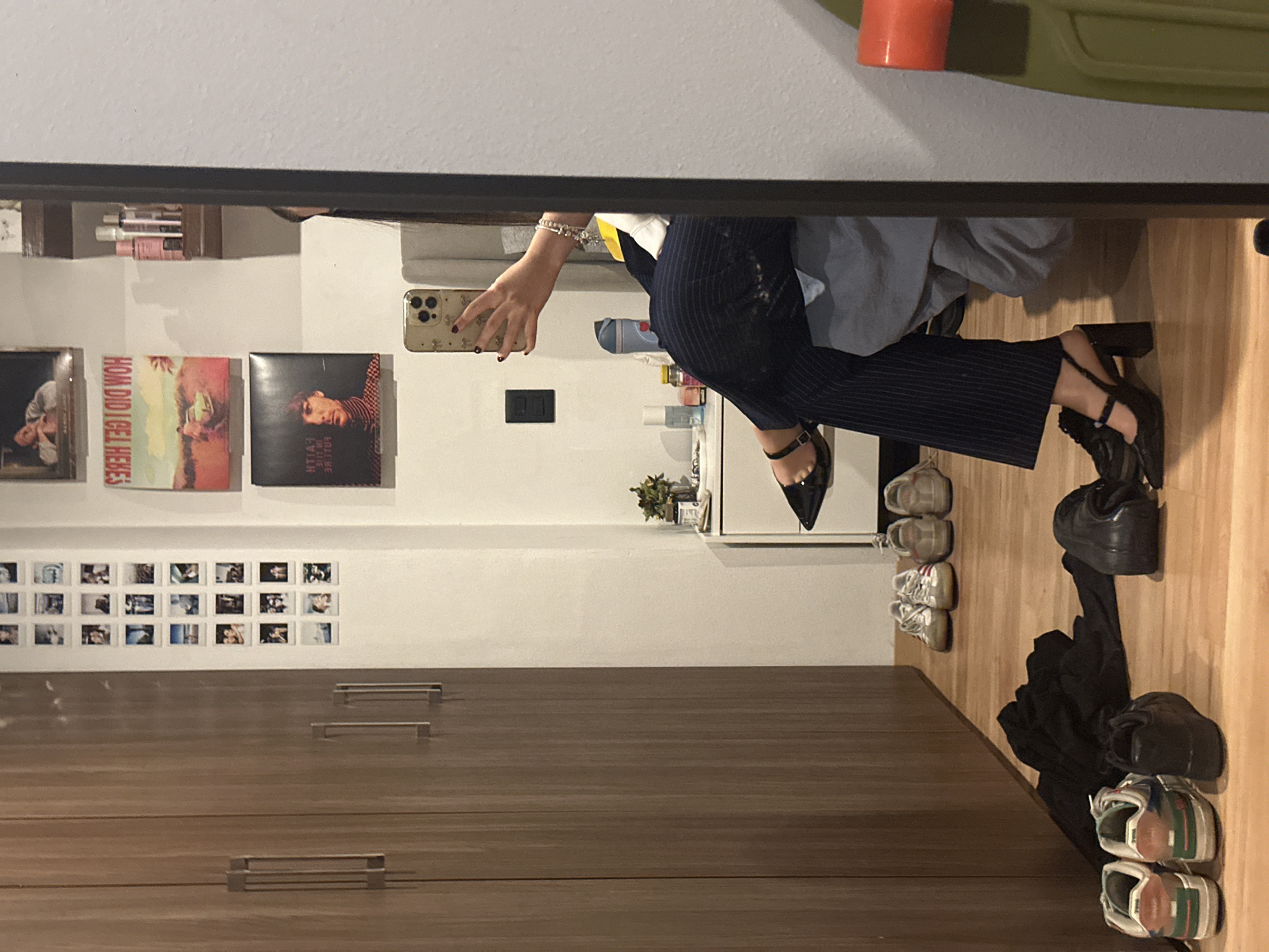 A person is taking a selfie in a mirror. The person is wearing a white shirt, blue jacket, navy striped pants, and black shoes with heels and ankle straps. The person is holding a gold iPhone with a decorative case. The mirror reflects a room with a white wall decorated with photographs, album covers, and a small plant. There is a wooden cabinet with metal handles on the lower part of the wall. On the floor, there are several pairs of shoes and a black garment. The location of the photo is a room, presumably a closet, in a location that is possibly Huixquilucan de Degollado, Mexico.