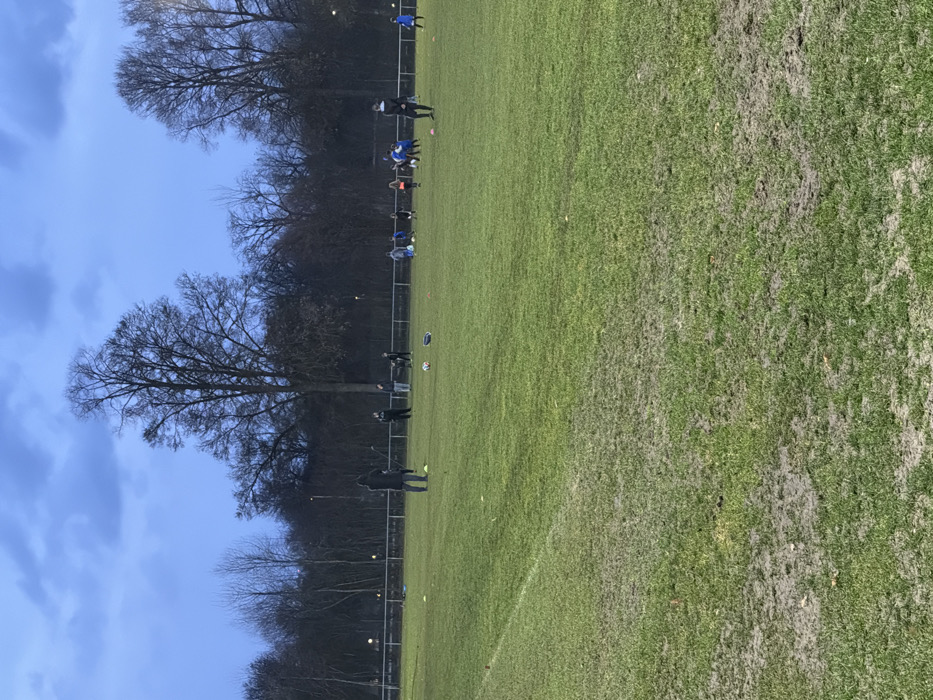 An outdoor grass sports field in Neu-Ulm, Germany, is depicted under an overcast, dark blue-grey sky, indicative of late afternoon or dusk. Approximately 15-20 individuals are present on the field, predominantly children and adolescents in athletic wear, alongside several adults, presumed to be coaches. They are distributed across the mid-ground and background, engaged in what appears to be a soccer training session, evidenced by multiple soccer balls and orange training cones on the ground. The field surface shows significant wear, with prominent muddy patches and sparse grass, particularly in the foreground. A faint white line marking is visible on the left. A metal fence runs across the background, separating the field from a dense line of tall, bare deciduous trees. Faint artificial lights are discernible through the trees.