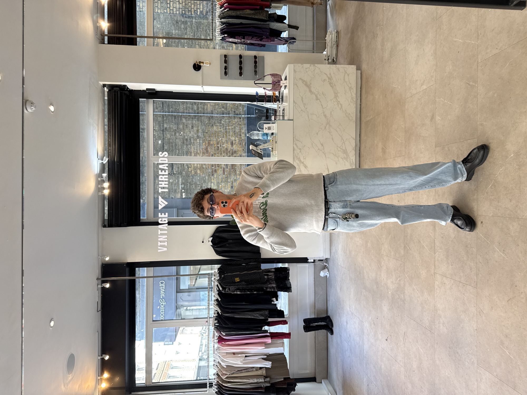 The photograph depicts an individual standing inside a retail store, likely a clothing boutique, in what appears to be London, United Kingdom. The store features racks of clothing displayed on hangers, with items ranging from jackets to shirts and pants. There is a counter with what seems like accessories for sale. The person in the image is taking a selfie with a phone, is wearing sunglasses, a light gray sweater, light blue jeans, and black shoes. The environment suggests a modern retail space. There is a sign for "Vintage Threads." The background shows building exteriors viewed through the storefront windows, indicative of an urban environment.