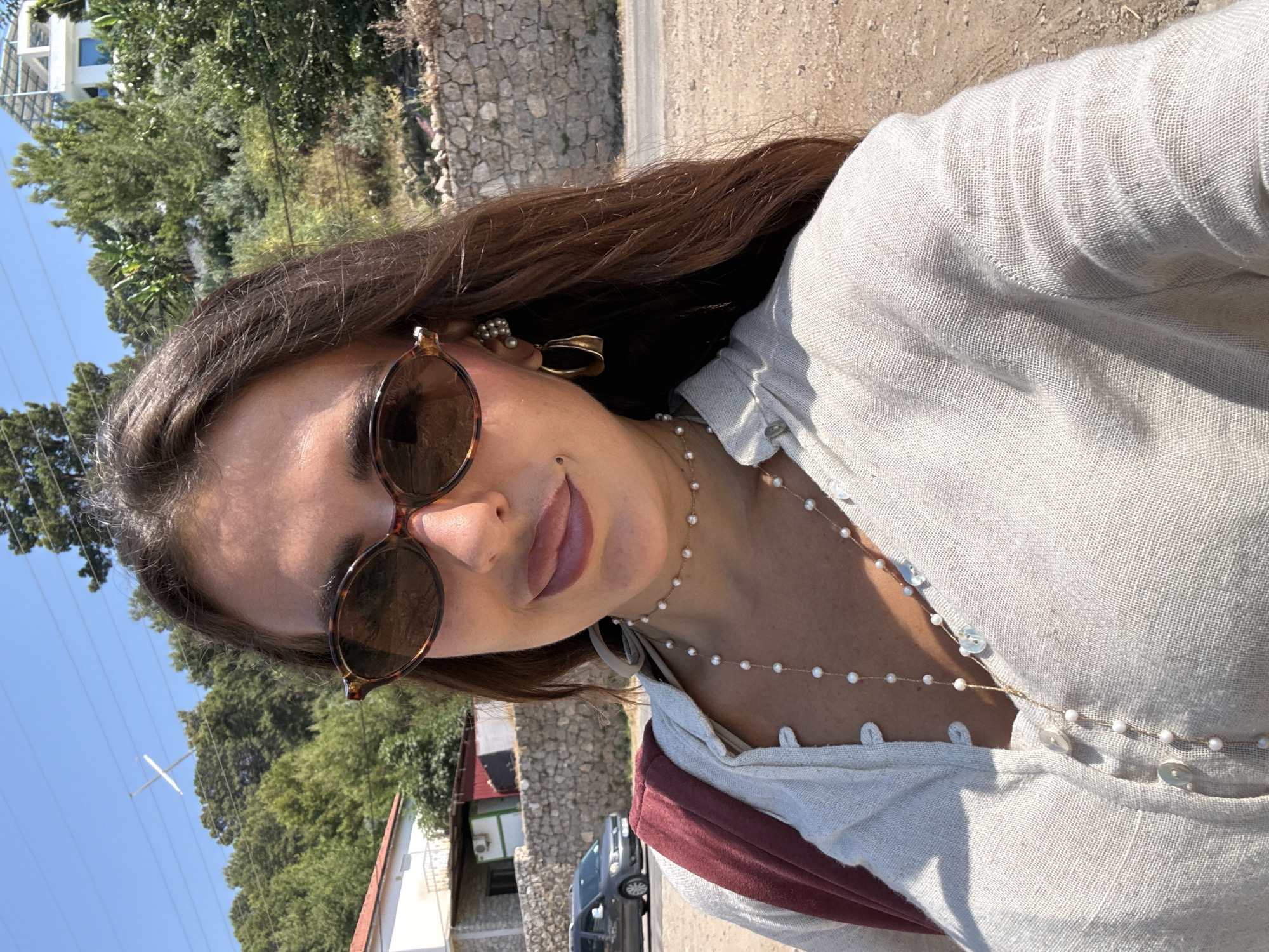 This is a close-up, vertically oriented selfie of a young woman with long, dark brown hair. She is wearing a light-colored, linen-like button-up shirt with a wine-colored garment underneath, visible at the neckline. She has on round, tortoise-shell sunglasses and delicate jewelry, including pearl earrings and a layered pearl necklace. Her lips are slightly parted, and she has a gentle smile. The sunlight casts shadows on her face, suggesting it is daytime.
The background is slightly blurred but shows a rocky wall, lush green trees, and a clear blue sky. A white building with a lattice-like structure is visible to the left. The overall impression is of a sunny day, likely outdoors in a warm climate. The image evokes a sense of casualness and enjoyment. The location is Fethiye, Turkey. There is no visible text in the image.