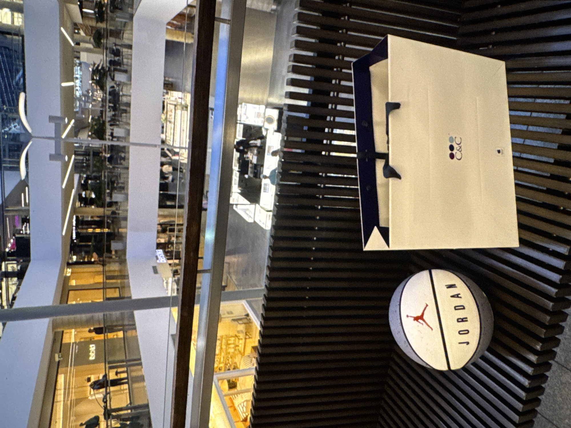The image depicts an indoor scene within a multi-level shopping mall, likely located in Kaunas, Lithuania, given the context. In the foreground, a basketball with the "JORDAN" logo and the Jumpman silhouette is placed on a dark wooden bench. To the right of the basketball is a light-colored shopping bag with the logo "C&C" and three colored circles.
The bench is positioned on a balcony or walkway overlooking a lower level of the mall. The background reveals the interior architecture of the mall, featuring walkways, storefronts with illuminated displays, escalators, and seating areas with tables and chairs. People are visible on different levels, some appearing to be shopping or dining. The overall lighting suggests it is daytime, with artificial lights illuminating the interior spaces. The environment appears clean and modern, consistent with a commercial shopping complex.