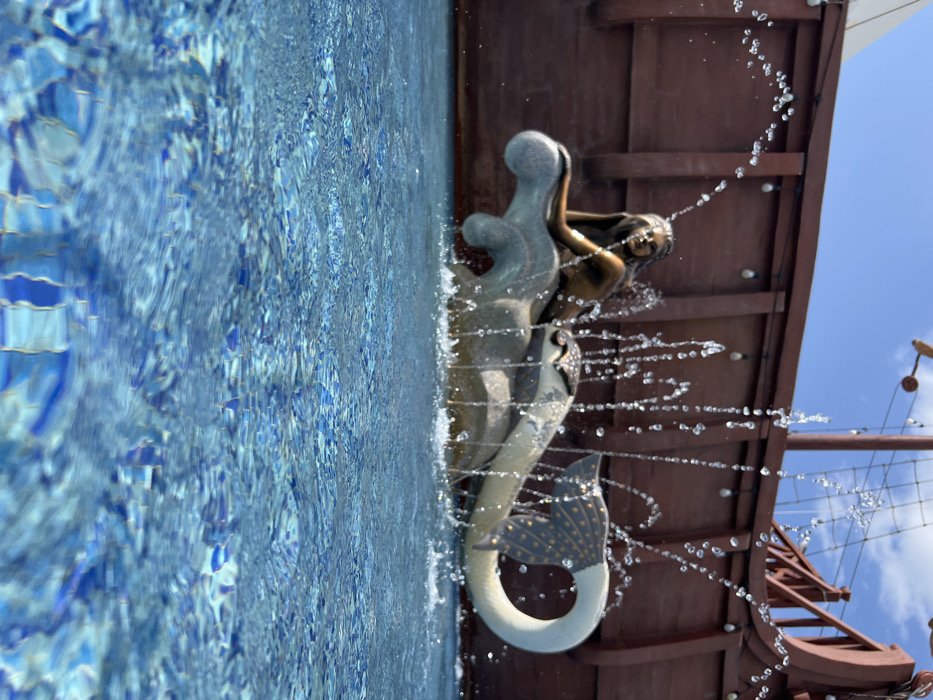 A mermaid fountain sculpture sits beside a pool with bright blue tiles. Water sprays from the mermaid. A portion of a ship’s structure is visible in the background, featuring dark wooden planks and nautical details. The sky, blue with light clouds, is also in view. The composition is unique, offering an eye-level perspective of the water feature.