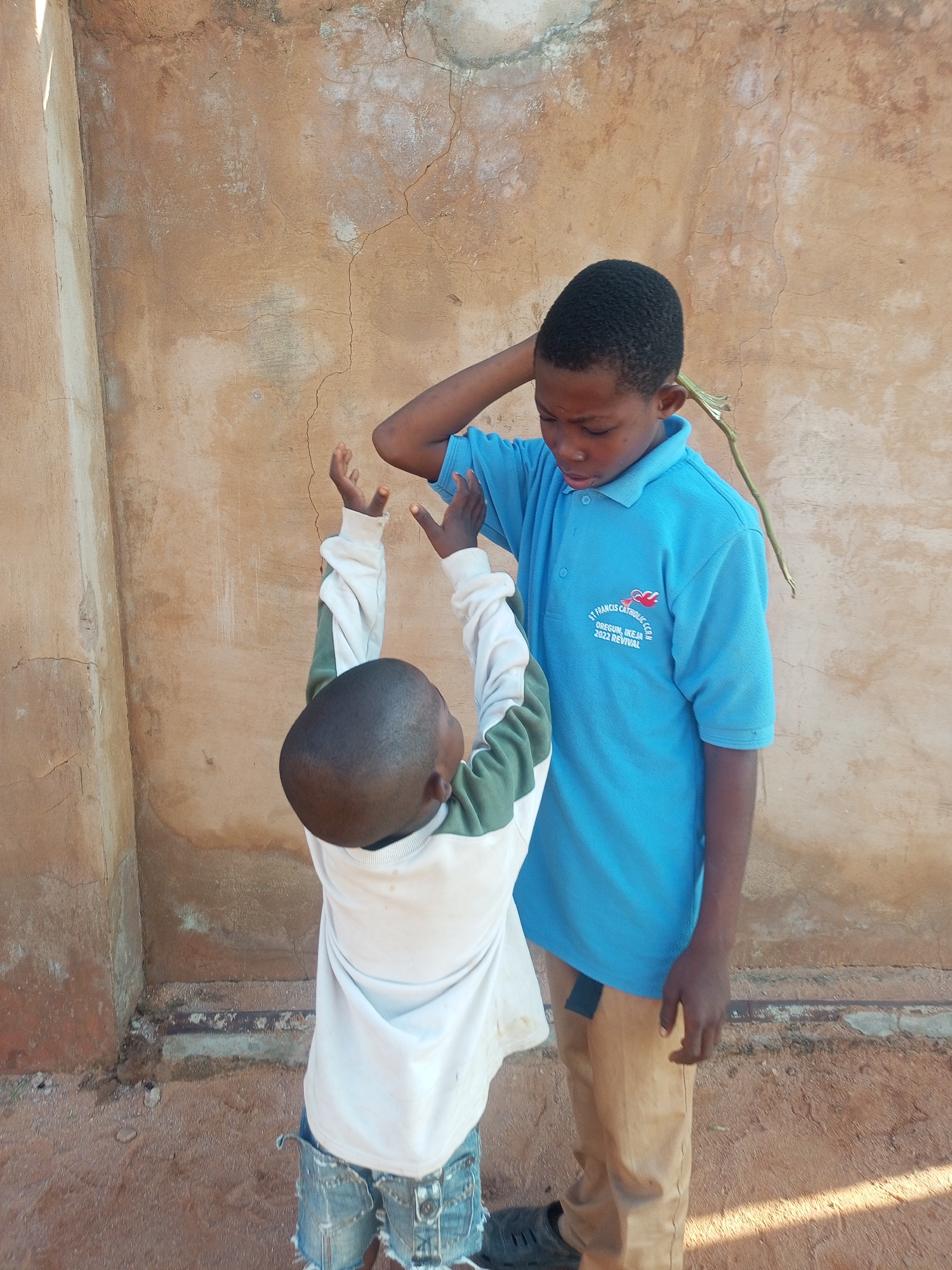 Two young Black boys stand outdoors against a textured, light brown wall. The boy on the right, who is taller, wears a bright blue polo shirt with a school logo and tan pants. A thin, green stick protrudes from behind his ear. His right arm is bent at the elbow with his hand resting on his head, while his left arm is lowered. The boy on the left, shorter and facing away from the camera, wears a white long-sleeved shirt with green accents on the sleeves and distressed denim shorts. His arms are raised towards the taller boy, with his hands open. The ground beneath them is reddish-brown dirt. The scene appears to be in Tempure, Nigeria.