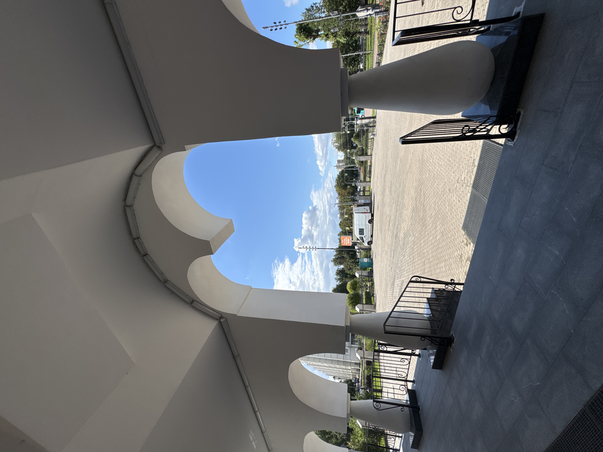 The image shows a view through a white arched colonnade, looking out onto a paved square under a bright blue sky with scattered clouds. In the background, there are trees, buildings, and a white van parked in the square. The architecture suggests a public building or institution. The scene appears to be in a city. The style of the architecture, specifically the white stone, arched openings and fluted columns, are characteristic of buildings in Saint Petersburg, Russia, likely a public institution such as a museum, government building or cultural center.