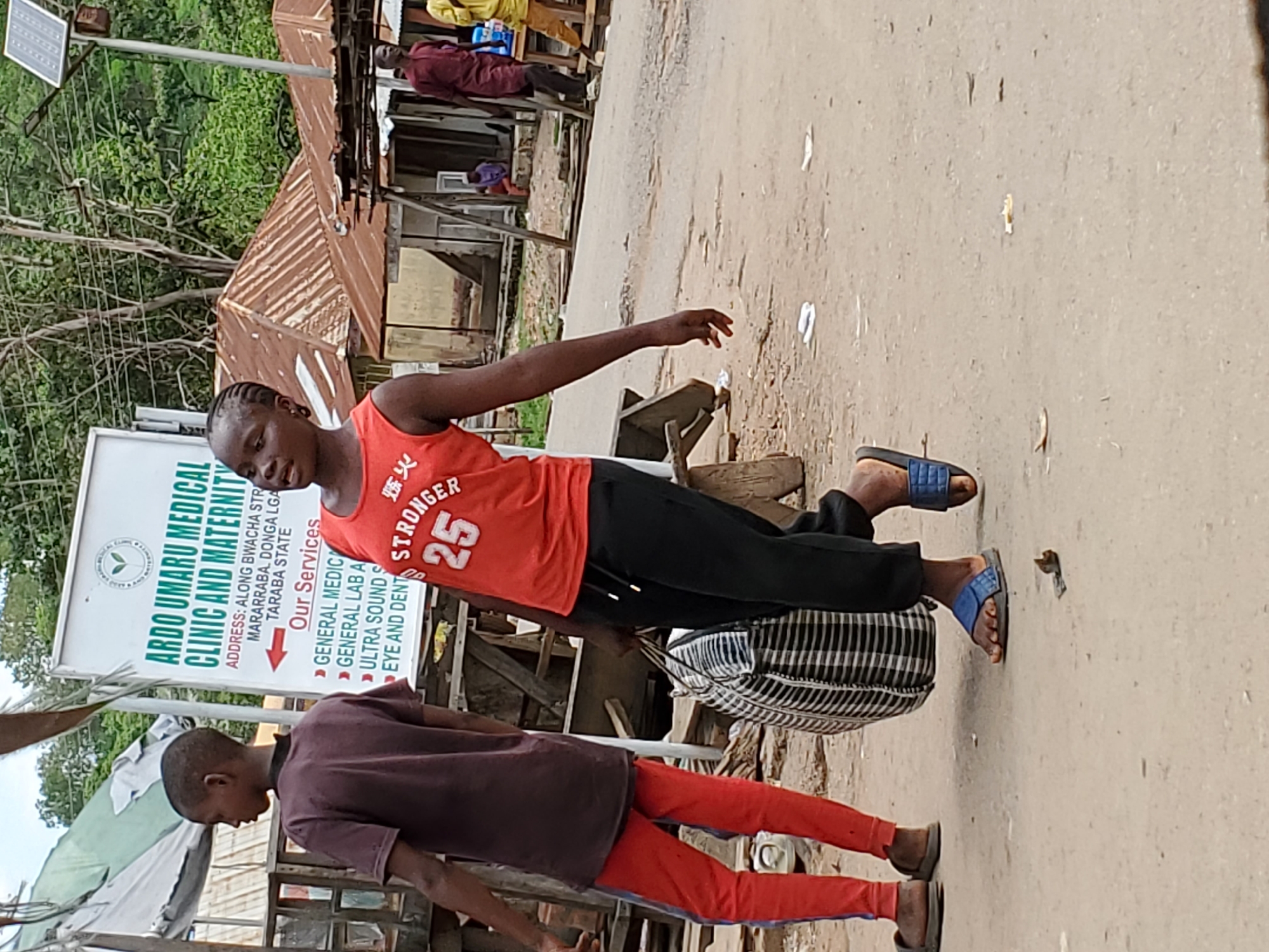In Mararaba, Nigeria, a street scene is captured, highlighting the Ardo Umaru Medical Clinic and Maternity. The clinic's sign advertises its services: general medicine, general lab, ultrasound, and eye/dental care. A young individual wearing an orange tank top with "Stronger 25" printed on it carries a checkered bag. Another person, dressed in a maroon shirt and red pants, stands nearby. The road is visible, and details of surrounding structures and vegetation are present.