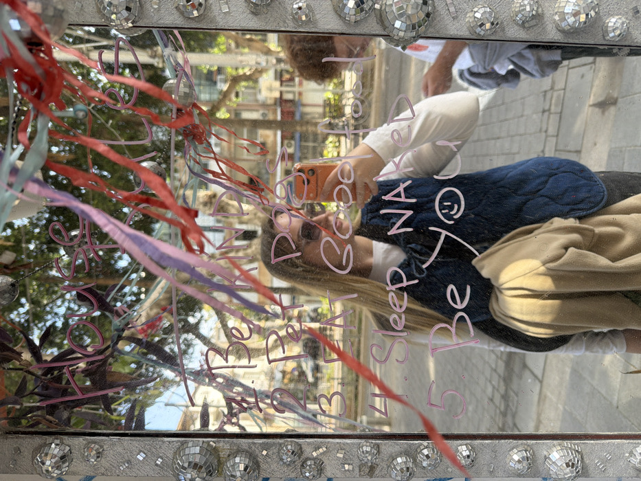 The image displays a reflection in a rectangular outdoor mirror or glass pane, framed with small disco ball tiles and adorned with numerous hanging ribbons in shades of red, pink, purple, and blue. Pink handwritten text on the surface reads "House" followed by an illegible word, and a numbered list: "1. Be KIND", "2. Pet Dogs", "3. Eat Good Food", "4. Sleep NAKED", and "5. Be You :)". A young woman with long blonde hair, wearing sunglasses, a white long-sleeved shirt, and a dark blue quilted vest, is prominently reflected, holding an orange smartphone as if taking the photograph. A beige garment hangs from her arm. To her right, a second individual with curly brown hair is partially reflected, looking downwards. The reflected background reveals an urban street scene in Tel-Aviv, Israel, featuring multi-story buildings with balconies, green trees, a paved sidewalk, and bright daylight, consistent with a public space or street.