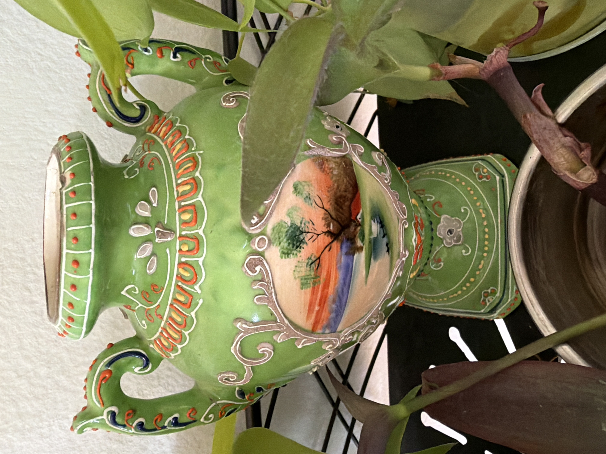 An ornate, green ceramic vase with two handles is positioned on a black shelf. The vase features intricate details including a painted landscape on the front, decorative patterns, and colored embellishments. The scene appears to be indoors, with a plain white wall in the background. The vase is surrounded by various green plants, some of which partially obscure the vase. A metallic pot is visible at the bottom of the image. There are no people or animals present. The location is unknown.