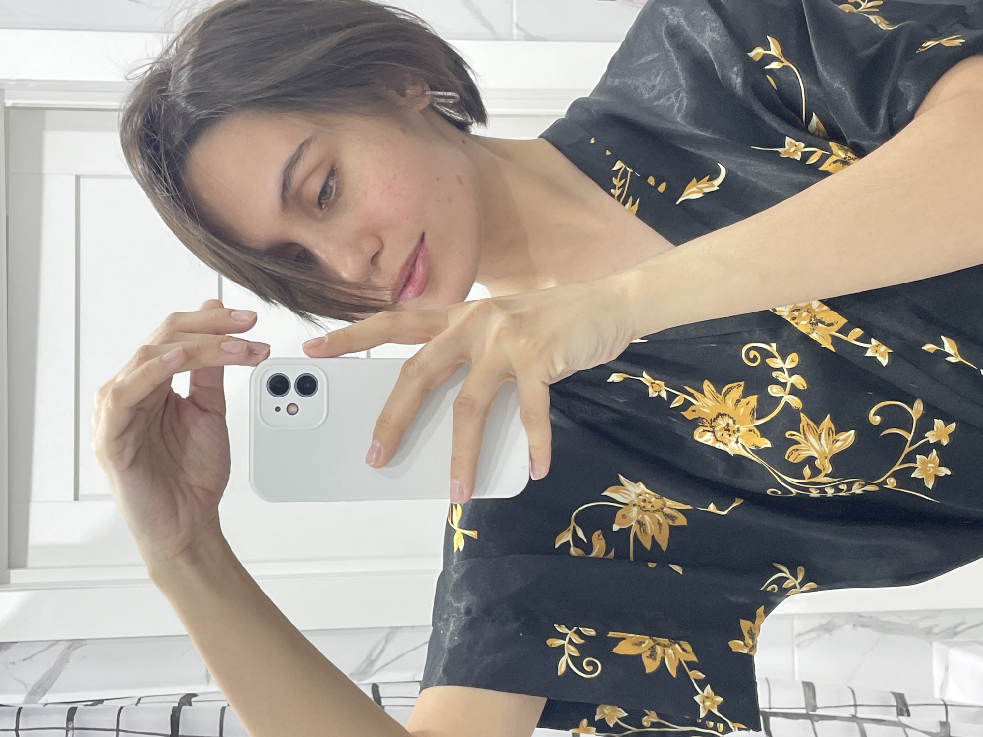 A person with short brown hair is holding a white smartphone and looking at it. The person is wearing a black robe with a gold floral pattern. The background appears to be a bathroom, with a white wall and a black and white checkered pattern at the bottom. The location is not discernable as Izhevsk, Russia, based solely on the image.