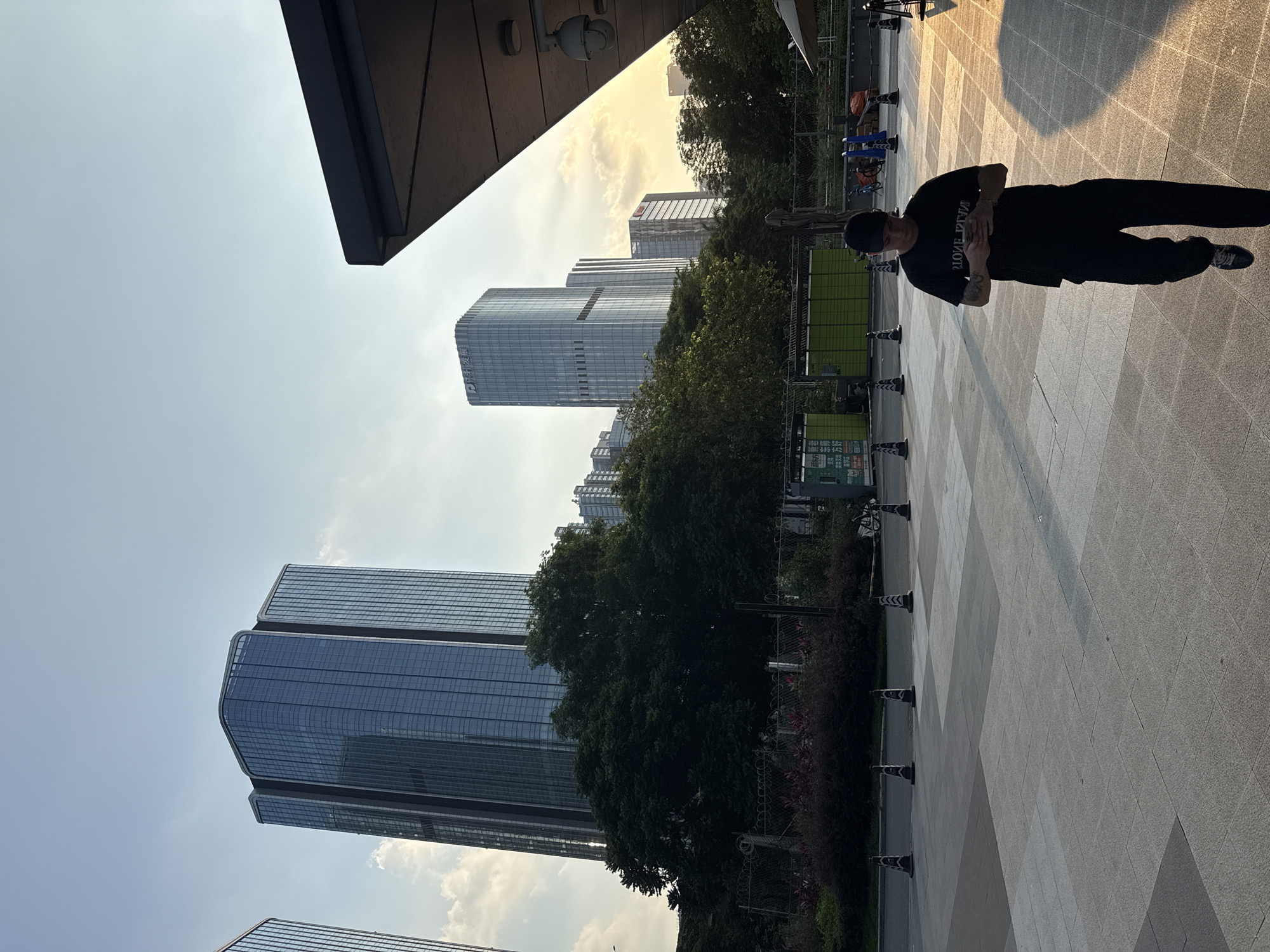 A slightly rotated eye-level shot reveals an urban landscape with modern architecture in Huangpu District, China. Tall glass-paneled buildings rise in the background, partially obscured by green trees. On the right, a man wearing a black t-shirt and pants is standing on a paved area with a grid pattern. Small black and white traffic cones run along the edge of the pavement near a line of shrubs. Overhead, a dark overhanging structure and a security camera are visible. The sky is clear with a hint of clouds around the building tops.