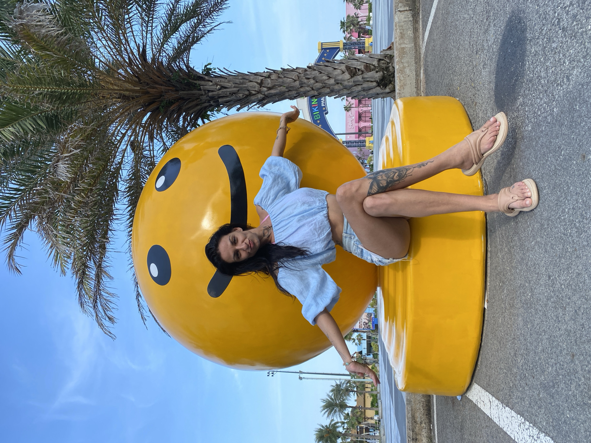 A woman with long, dark hair sits on a large yellow smiley face sculpture in a paved outdoor area. She is wearing a light blue off-the-shoulder crop top and denim shorts. Her left arm is extended outwards, and her right arm rests on her knee. She has a tattoo on her left thigh. A palm tree is visible behind her. In the background, there is a bright blue sky with some clouds, palm trees, and buildings with colorful signage, including "BIKINI PHU QUOC". The location is identified as Tien Thanh Ward, Vietnam.